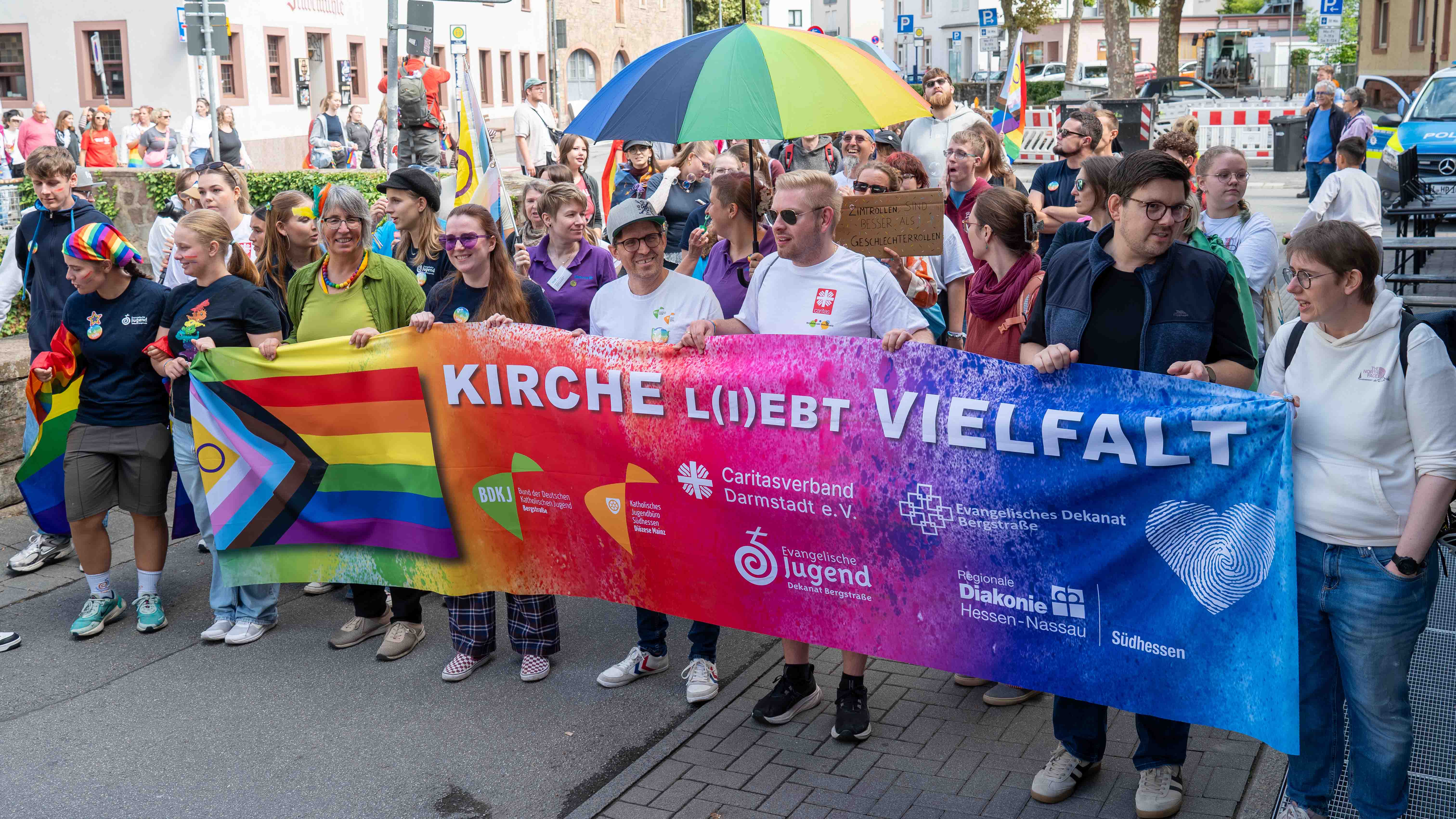 Menschen beim CSD halten ein buntes Banner mit der Aufschrift „Kirche liebt Vielfalt“ und eine Progress-Pride-Flagge.