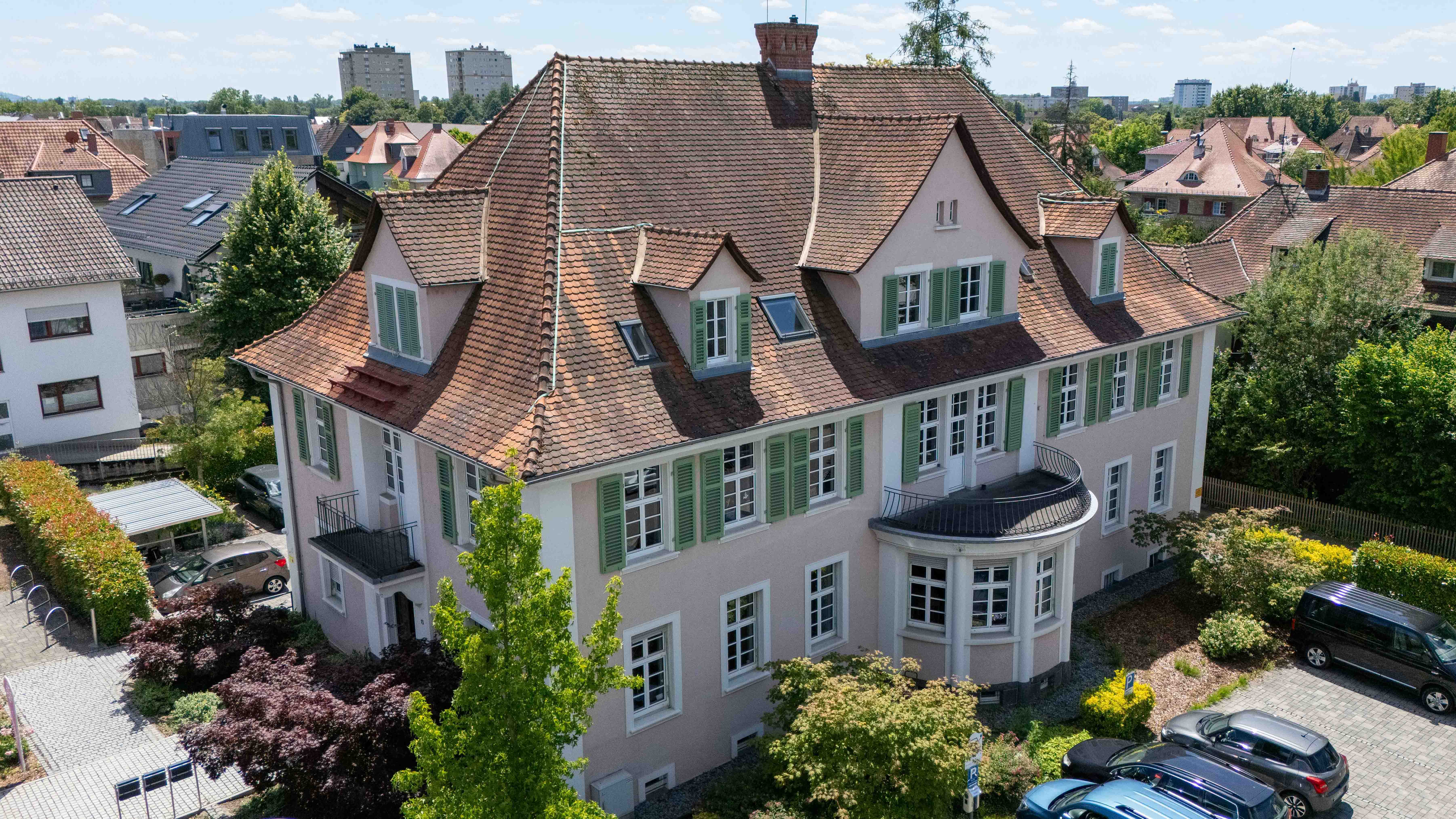 Großes mehrstöckiges Wohnhaus mit rotem Ziegeldach, hellen Fassaden und grünen Fensterläden, umgeben von Bäumen, Gärten und parkenden Autos.