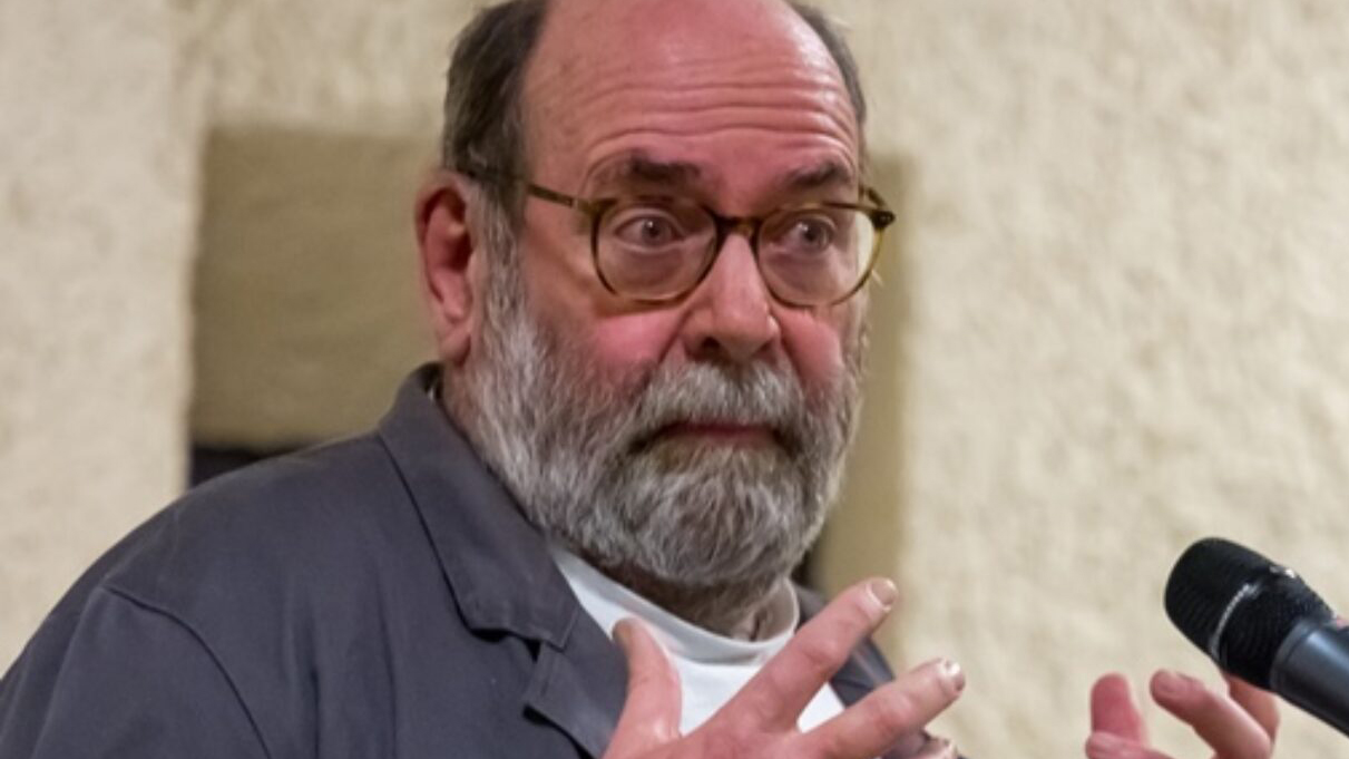Älterer Mann mit Brille und grauem Bart spricht an einem Mikrofon und gestikuliert mit beiden Händen; sein Blick ist aufmerksam zur Seite gerichtet.