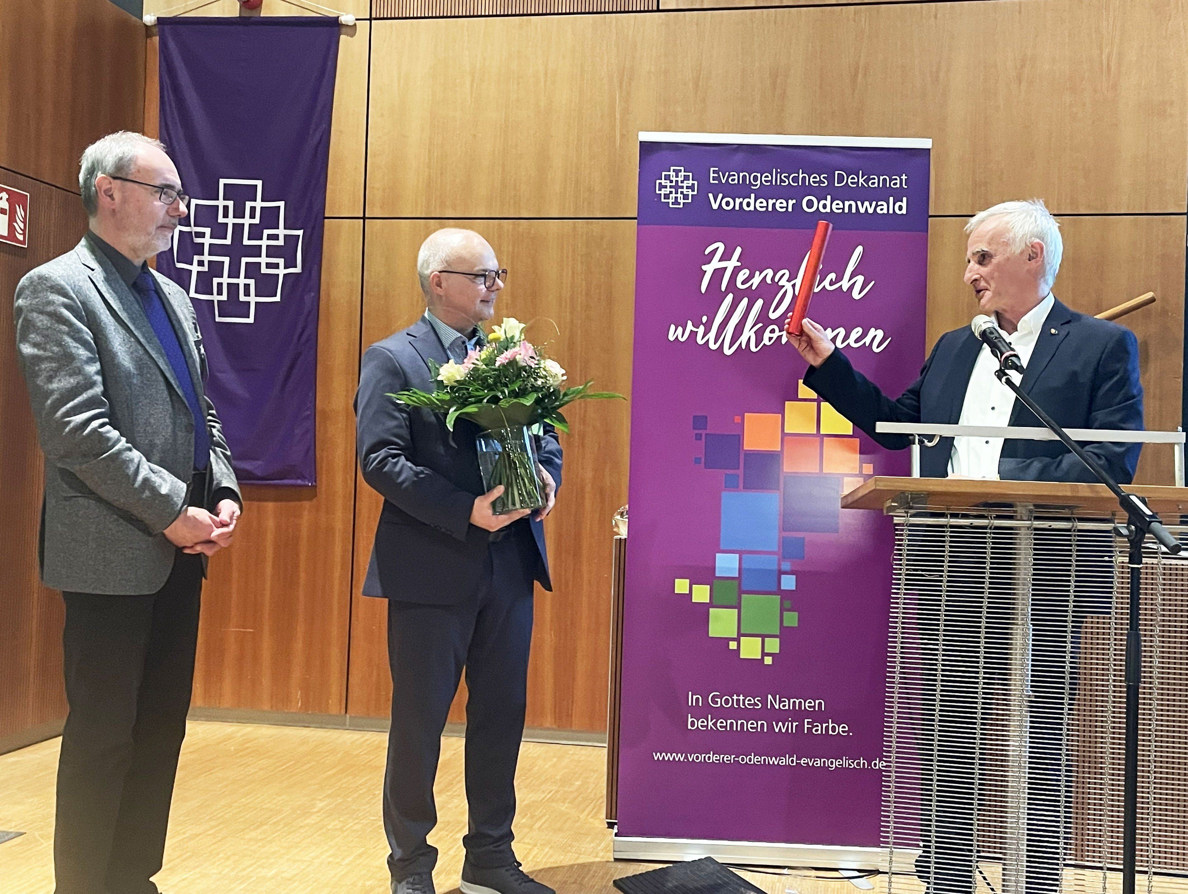 Neuer Dekan im Vorderen Odenwald Drei Männer, einer bekommt Blumen.