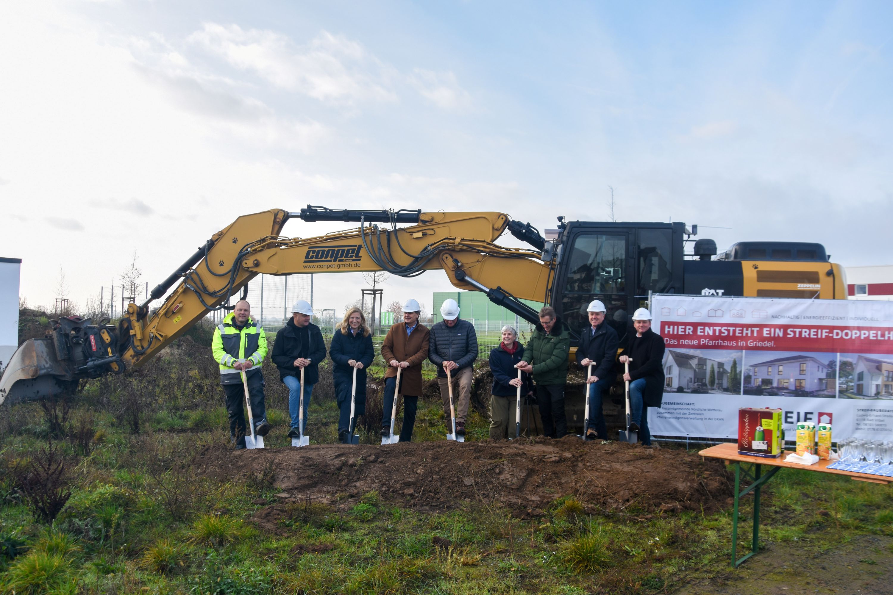 Menschen mit Schaufeln in der Hand vor einem Bagger auf dem Bauplatz des neuen Pfarrhauses.