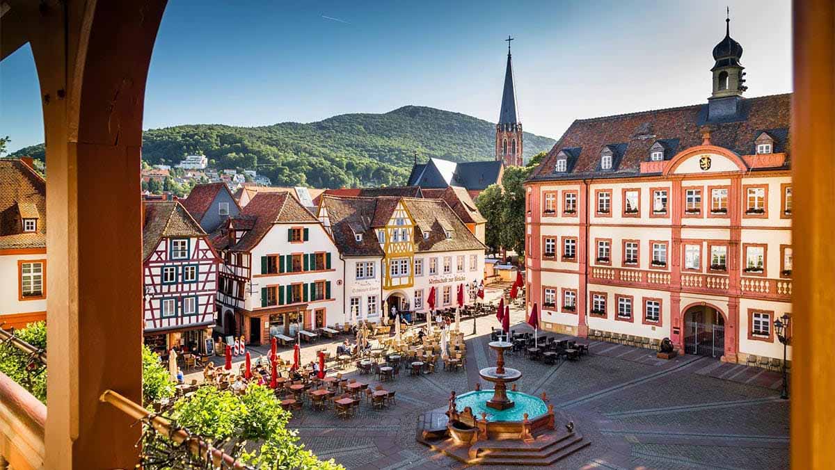 Historischer Marktplatz mit Fachwerkhäusern, Rathaus, Straßencafés und Brunnen; im Hintergrund eine Kirche und bewaldete Hügel.