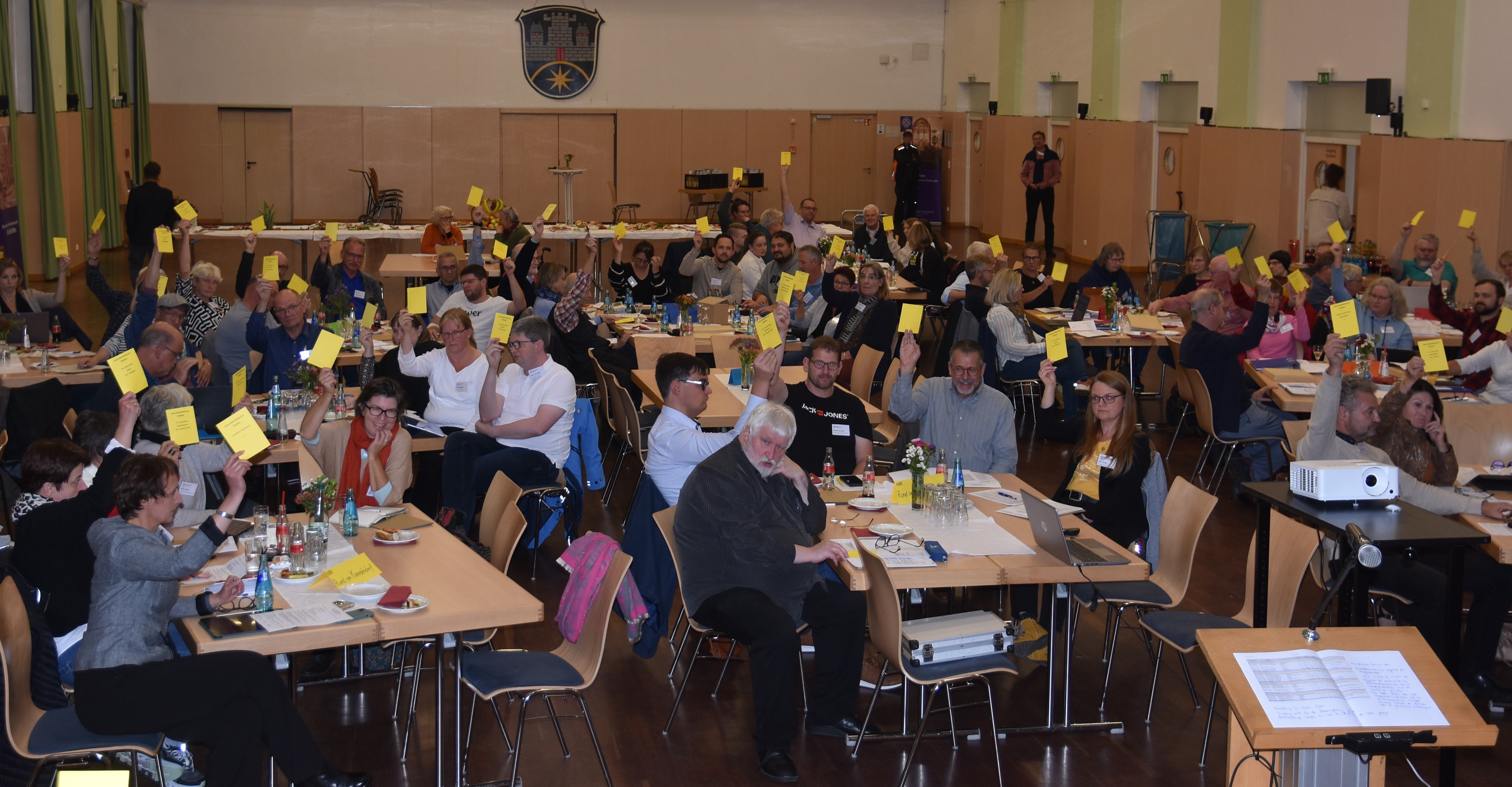 Blick in den Saal des Bürgerhauses Nidda, die Synodalen halten gelbe Stimmkarten in die Höhe und stimmen ab.