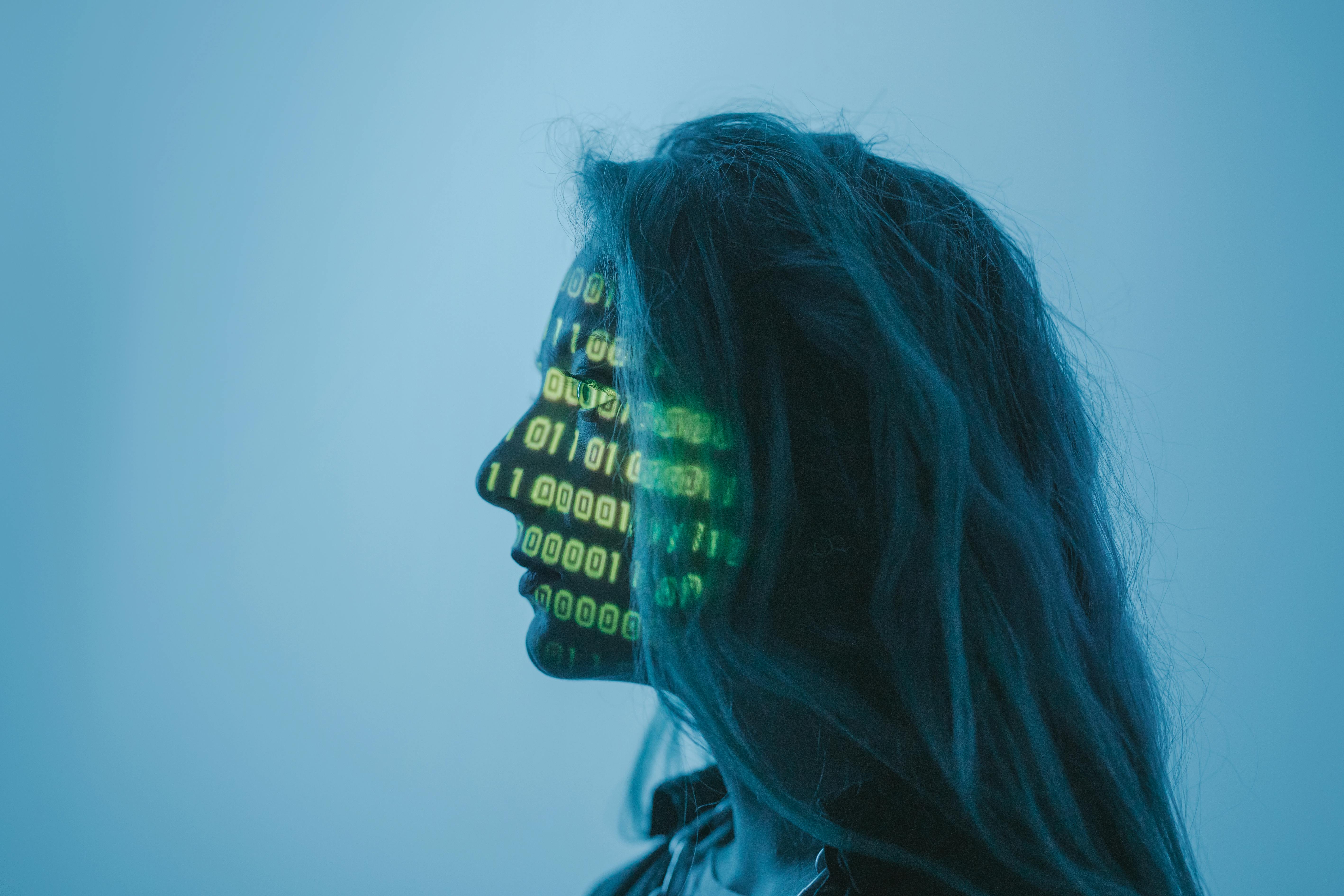Das Bild zeigt das Profil einer Frau, auf dessen Gesicht und Hals ein Muster aus digitalen Codes oder Daten projiziert wird. Die Farben sind überwiegend in Blau- und Grüntönen gehalten, was dem Bild eine futuristische oder technologische Atmosphäre verleiht. Es könnte eine Darstellung der Verbindung von Mensch und Technologie, künstlicher Intelligenz oder digitaler Identität sein.