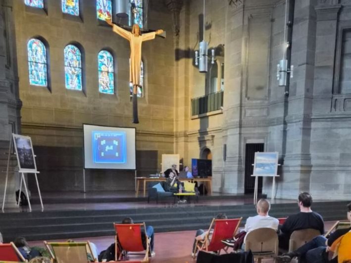 Menschen sitzen auf Liegestühlen im Kirchenraum im Hintergrund hängt Jesus am Kreuz über dem Altar.