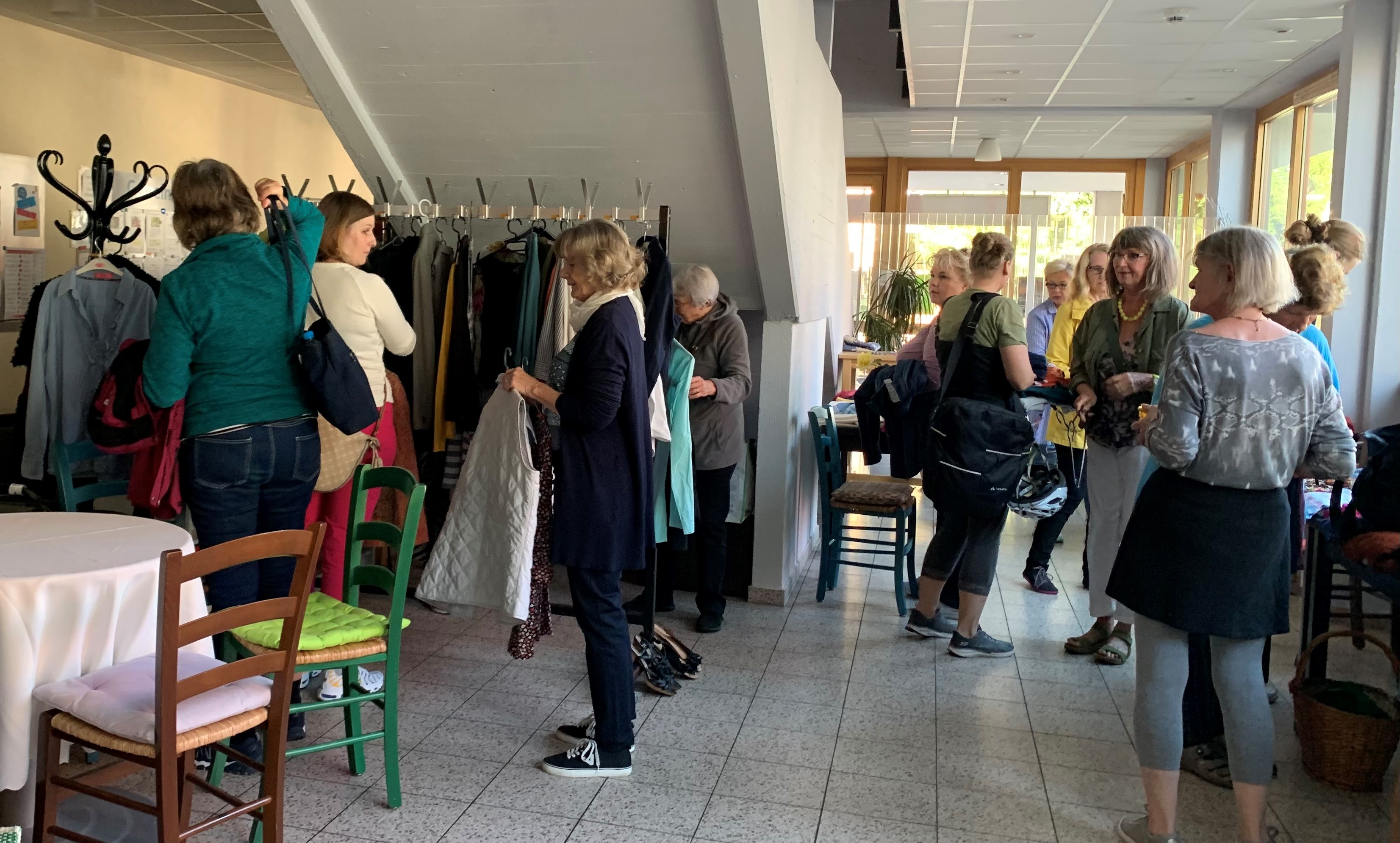 Das Bild zeigt eine Gruppe von Menschen in einem Gebäude. Einige durchstöbern Mäntel und Kleidungsstücke, die an Kleiderständern hängen. Andere unterhalten sich oder ordnen Gegenstände im Raum.