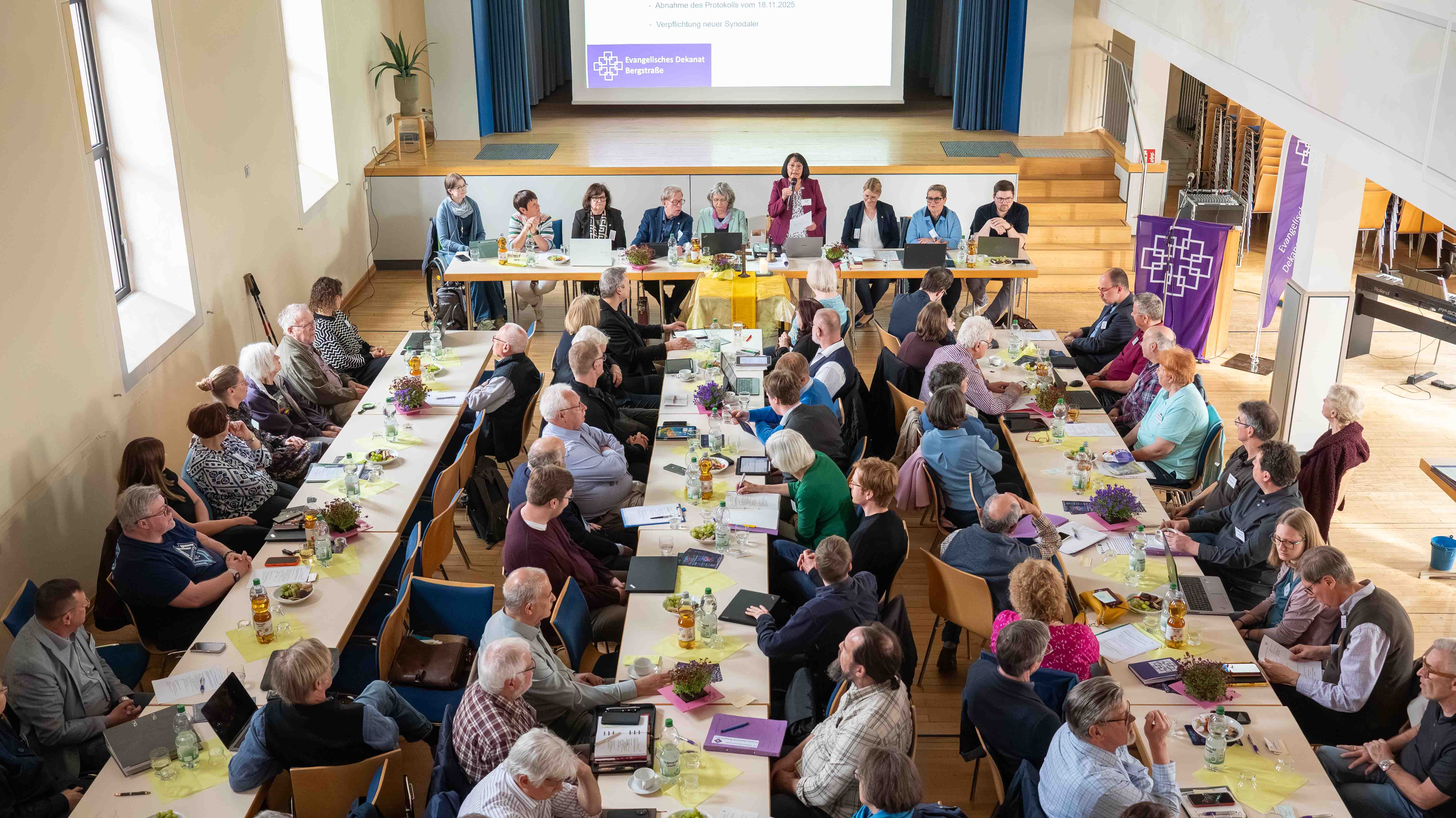 Großer Versammlungssaal mit mehreren Tischreihen, an denen zahlreiche Menschen sitzen und an einer Sitzung teilnehmen. Vorn auf einer Bühne sitzt ein Podium mit mehreren Personen; eine Frau spricht in ein Mikrofon. Im Hintergrund ist eine Präsentationsfläche mit dem Logo des Evangelischen Dekanats Bergstraße zu sehen. Die Teilnehmenden hören zu, diskutieren und arbeiten mit Unterlagen und Laptops.