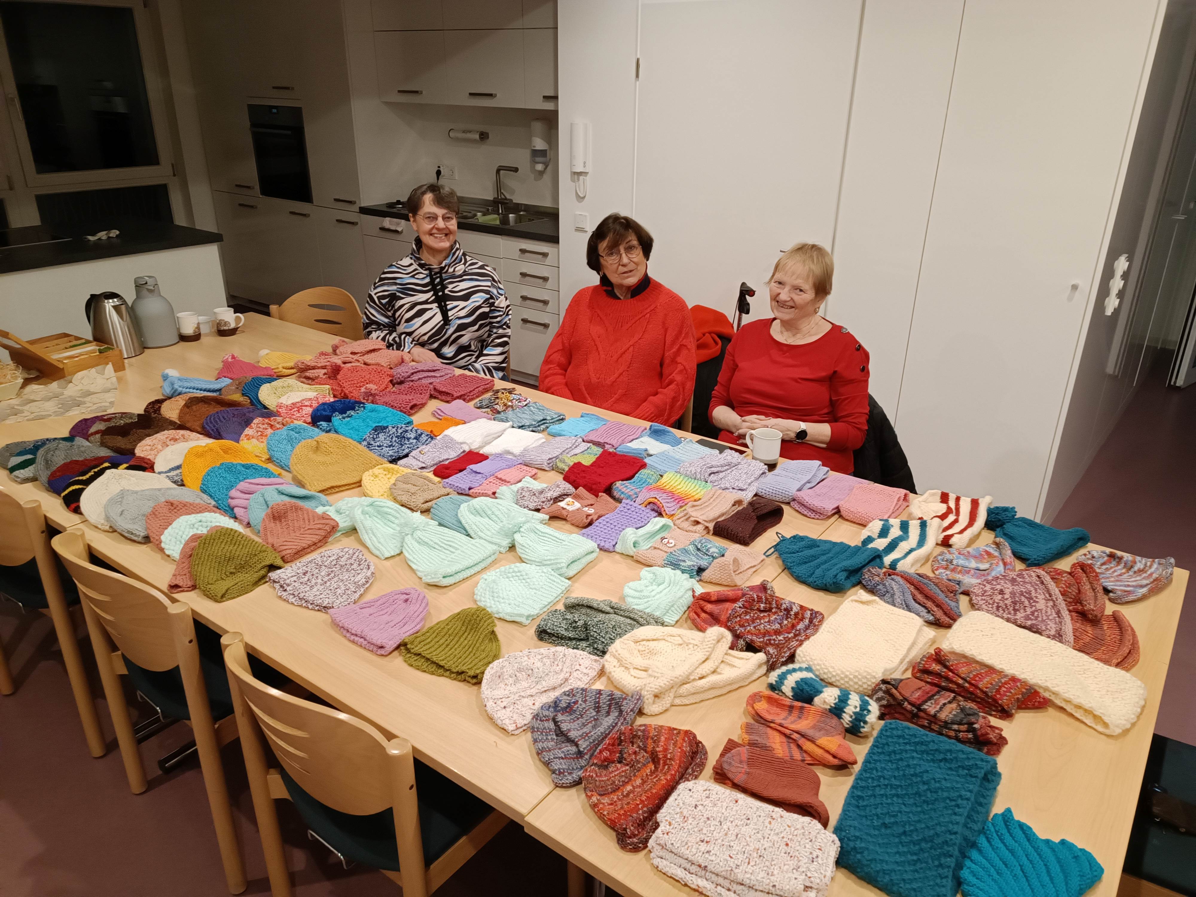 Strickartikel (Schals, Mützen, Handschuhe ) für Kinder liegen auf einem Tisch. Am Tisch sitzen 3 Frauen