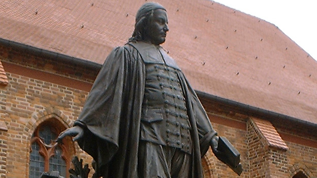 Bronzestatue eines Mannes in historischer Kleidung mit Buch in der Hand vor einer Backsteinkirche.