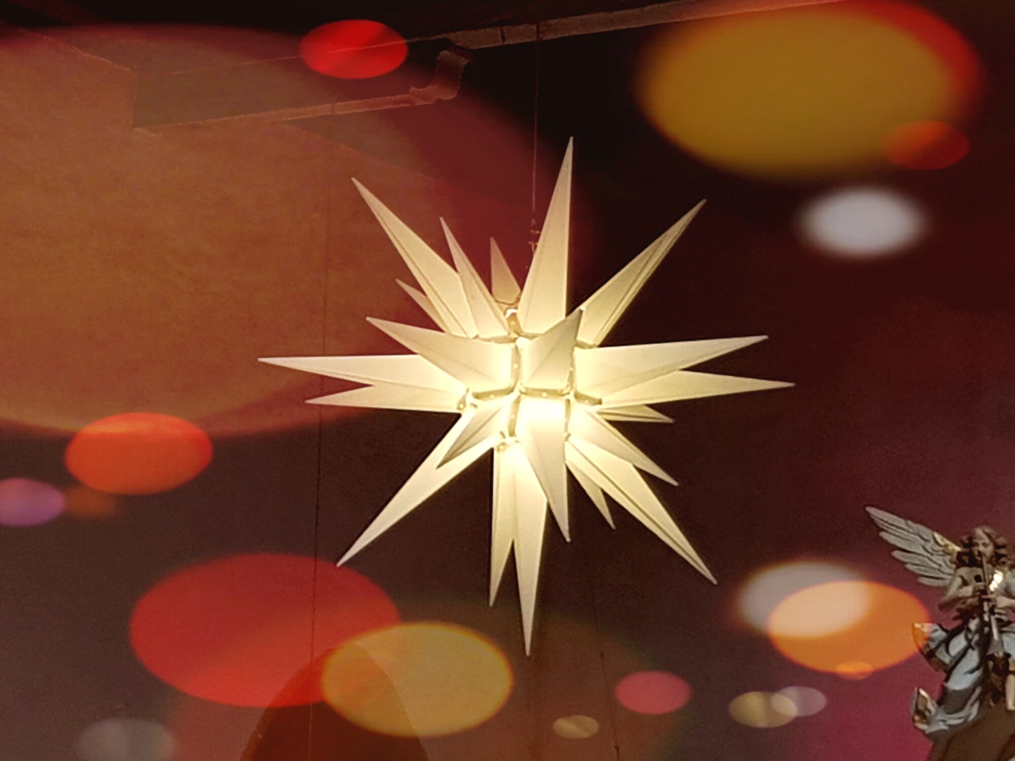 Das Bild zeigt einen weißen Herrnhuter Stern mit bunten Lichtpunkten an der Kirchendecke. In der rechten unteren Ecke ist ein Engel zu sehen.