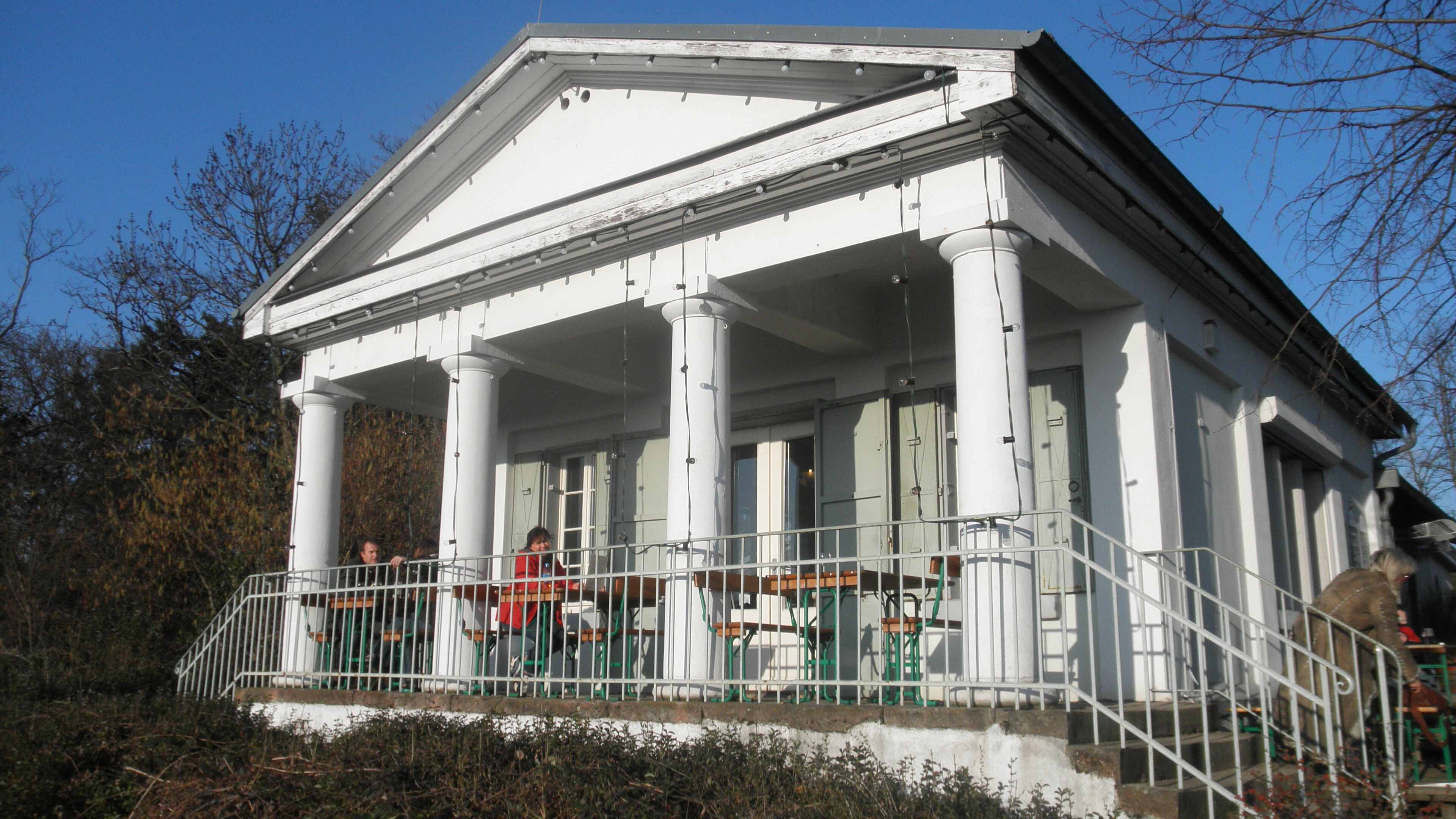 Weißes kleines Gebäude mit Säulen und Terrasse; zwei Personen sitzen draußen an Tischen, eine weitere Person geht eine Treppe hinauf, bei sonnigem Wetter.
