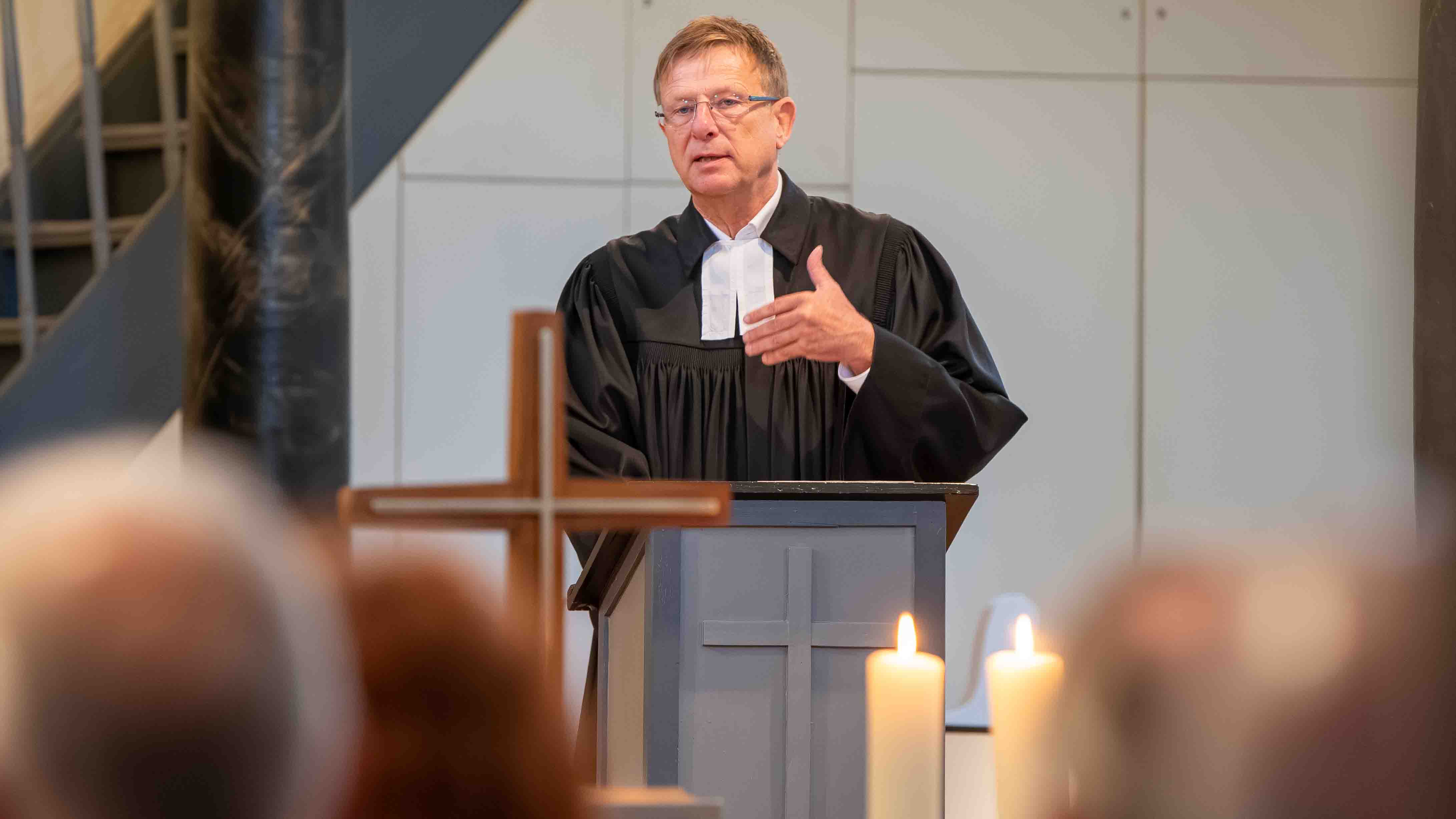 Pfarrer im schwarzen Talar spricht gestikulierend an einem Pult mit Kreuz; im Vordergrund sind ein Holzkreuz, brennende Kerzen und Gäste zu sehen.