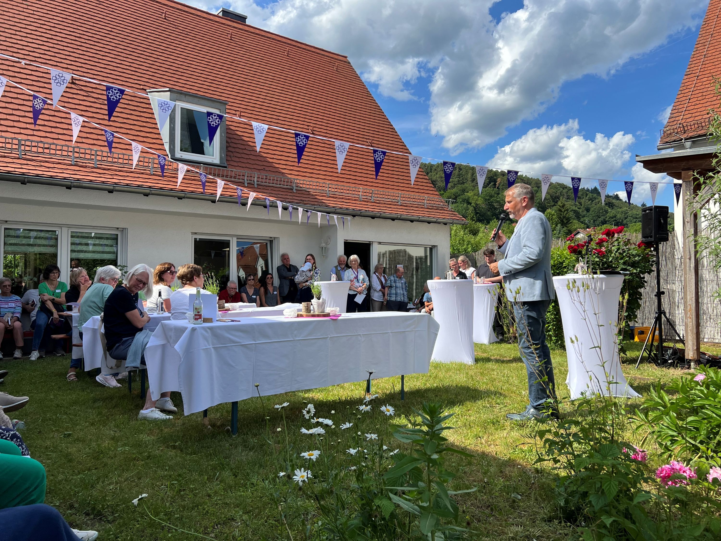 Ein Mann spricht in einem festlich geschmückten Garten in ein Mikrofon. In dem Garten stehen Menschen, andere sitzen an Tischen