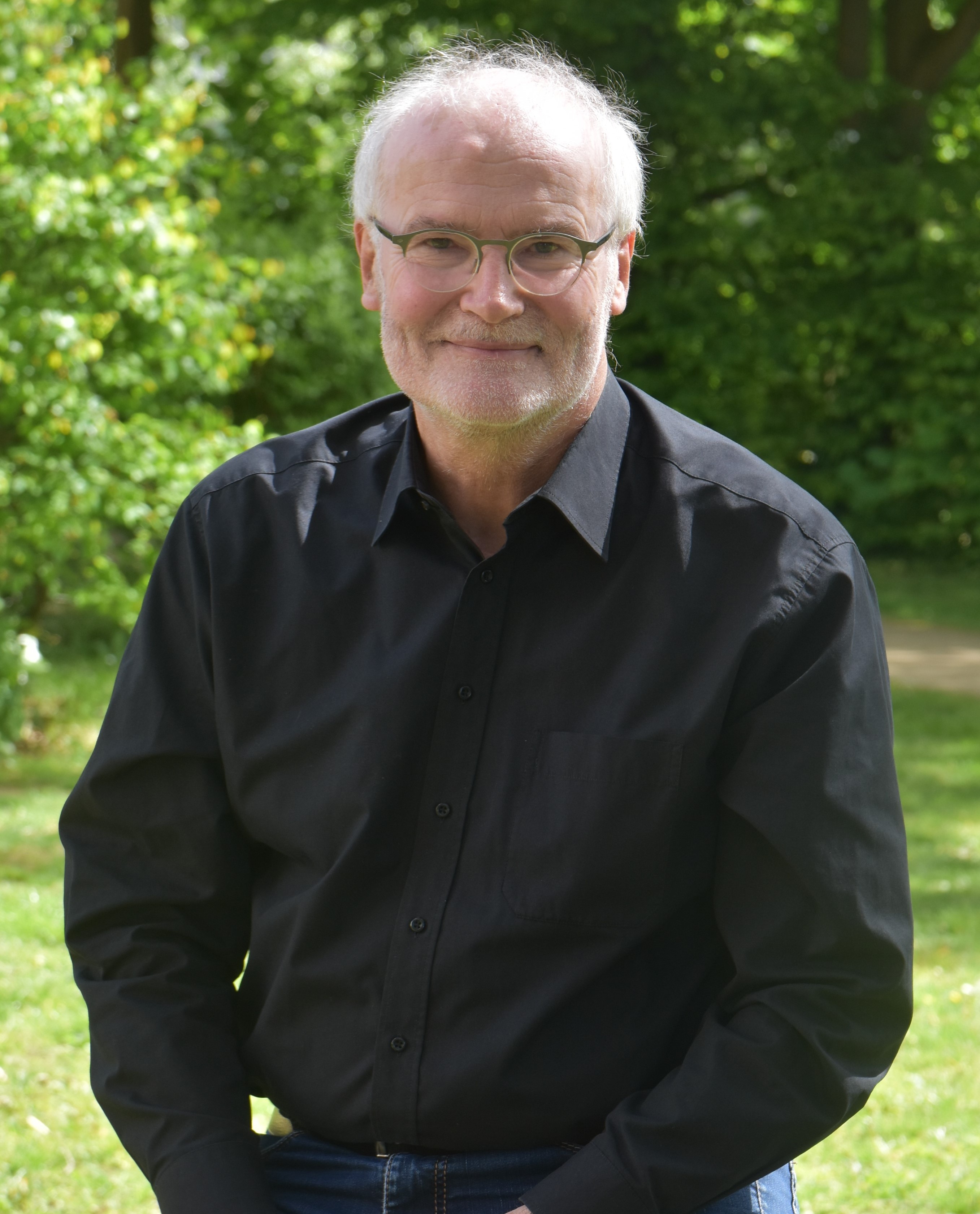 Rainer Böhm, aufgenommen unter freiem Himmel, trägt ein schwarzes Hemd und lächelt in die Kamera.