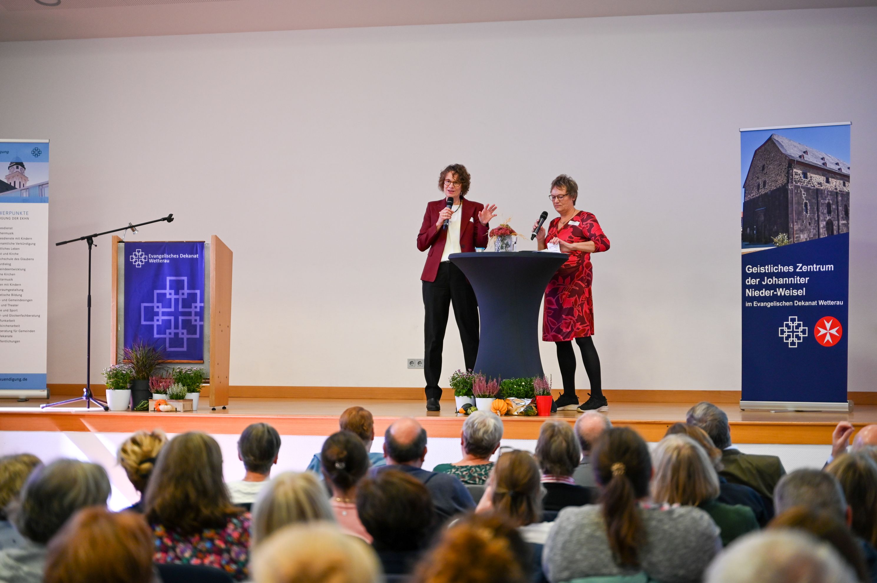 Zwei Frauen stehen mit Mikrofonen an einem Stehtisch auf einer Bühne und sprechen zum Publikum, das im Vordergrund von hinten zu sehen ist.