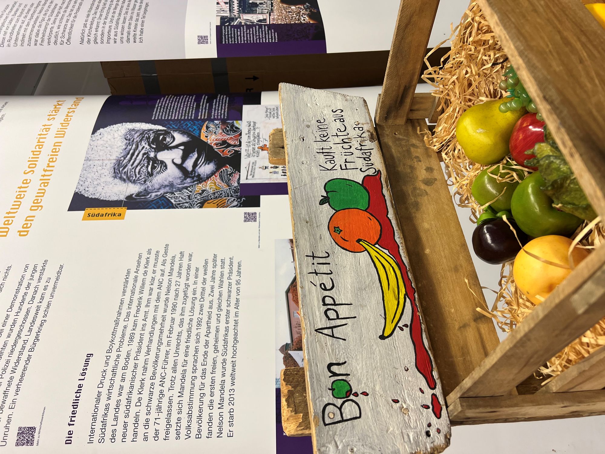Ausschnitt aus einer Ausstellung. Es ist eine Informationstafel zu sehen, darauf ist Nelson Mandela zu erkennen. Im Vordergrund steht eine Obstkiste.
