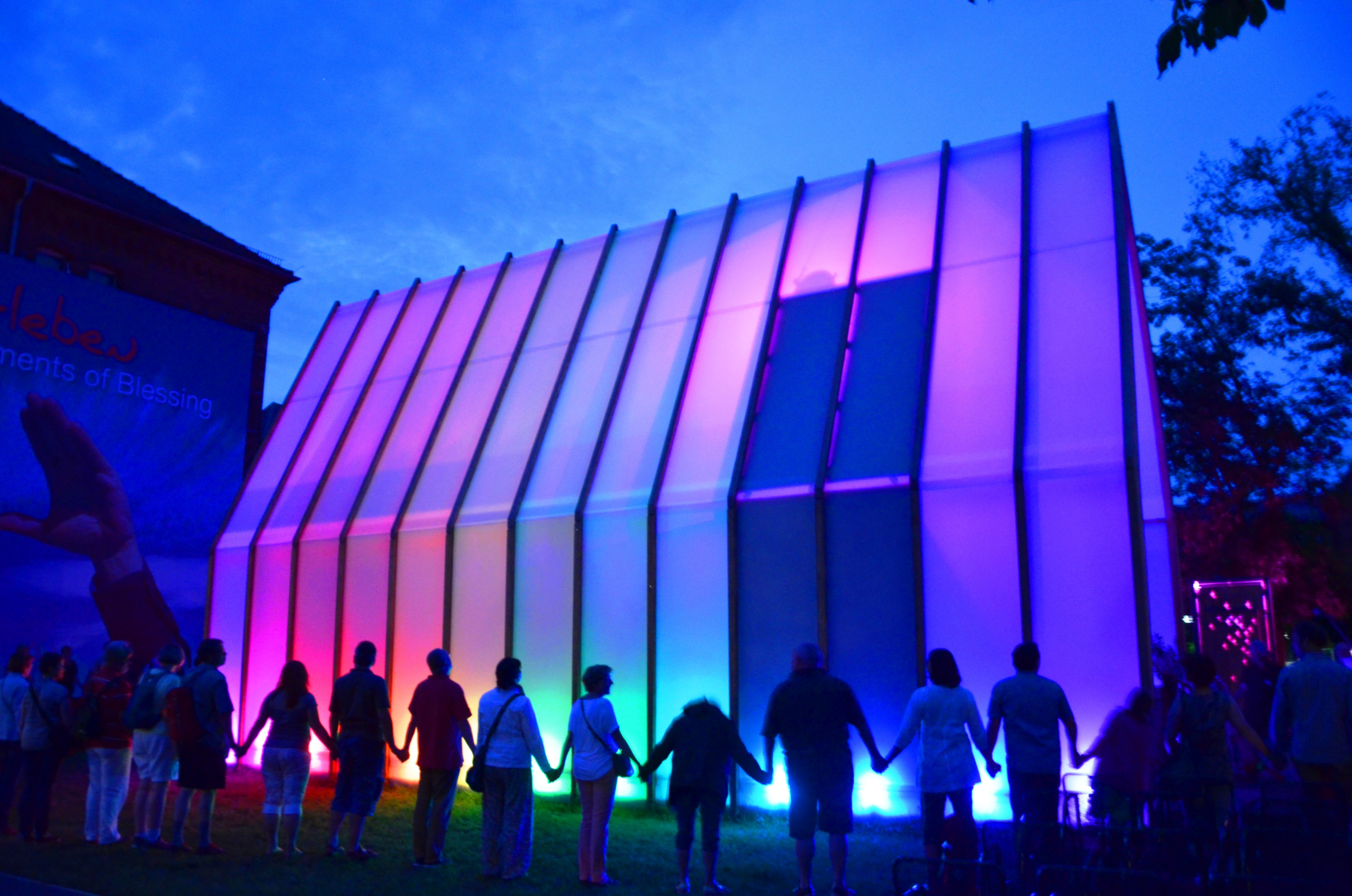 Die Lichtkirche, aufgenommen bei Dunkelheit, erstrahlt in den Farben des Regenbogens. Um die Kirche herum stehen Menschen, die sich an den Händen fassen.