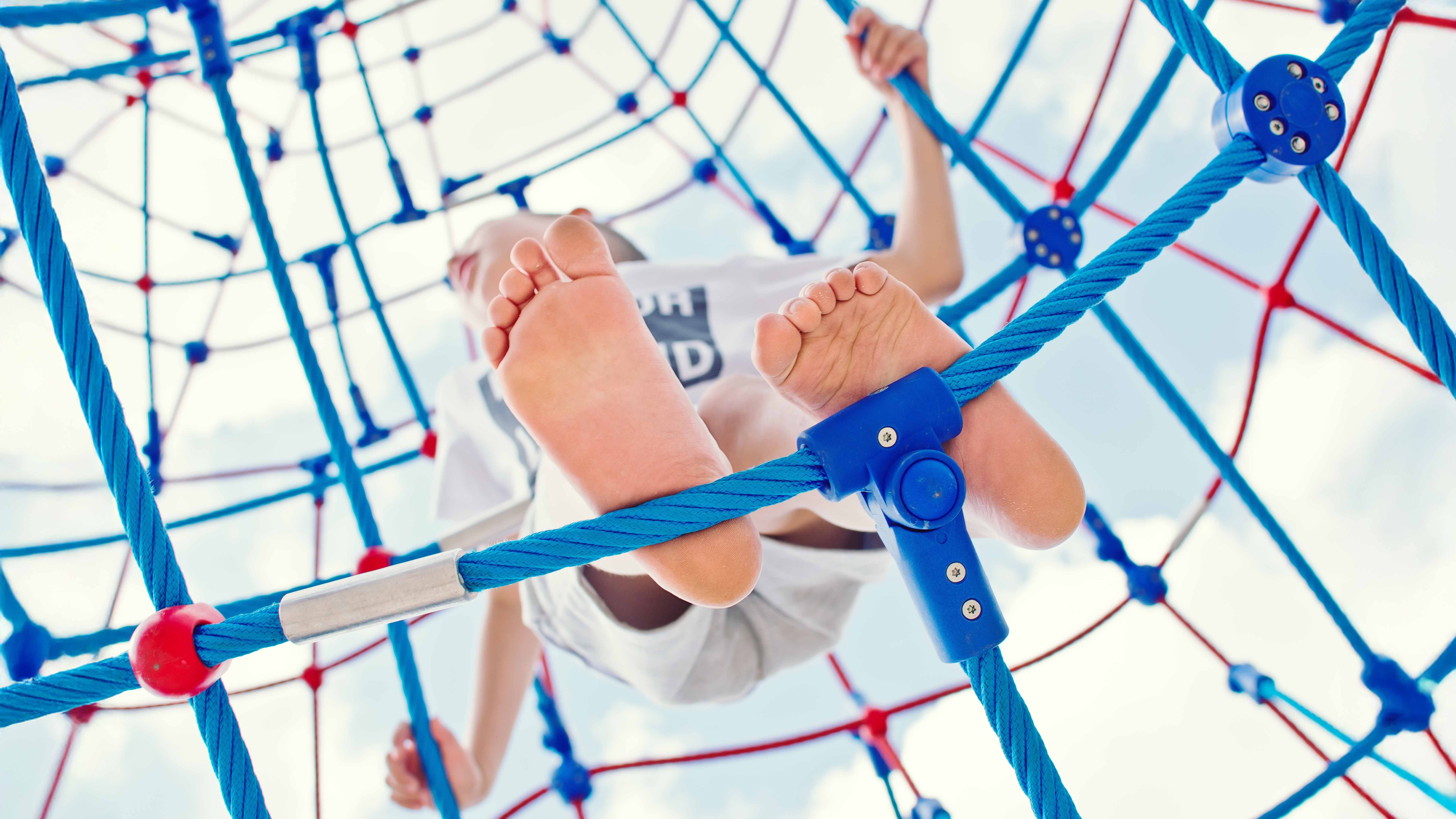 Ein Kind bewegt sich barfuß in einem großmaschigen Kletternetz aus blauen und roten Seilen auf einem Spielplatz. Die Aufnahme zeigt die Fußsohlen und Teile des Körpers aus einer Perspektive von unten, während das Kind sich am Netz festhält. Der Himmel ist im Hintergrund sichtbar.