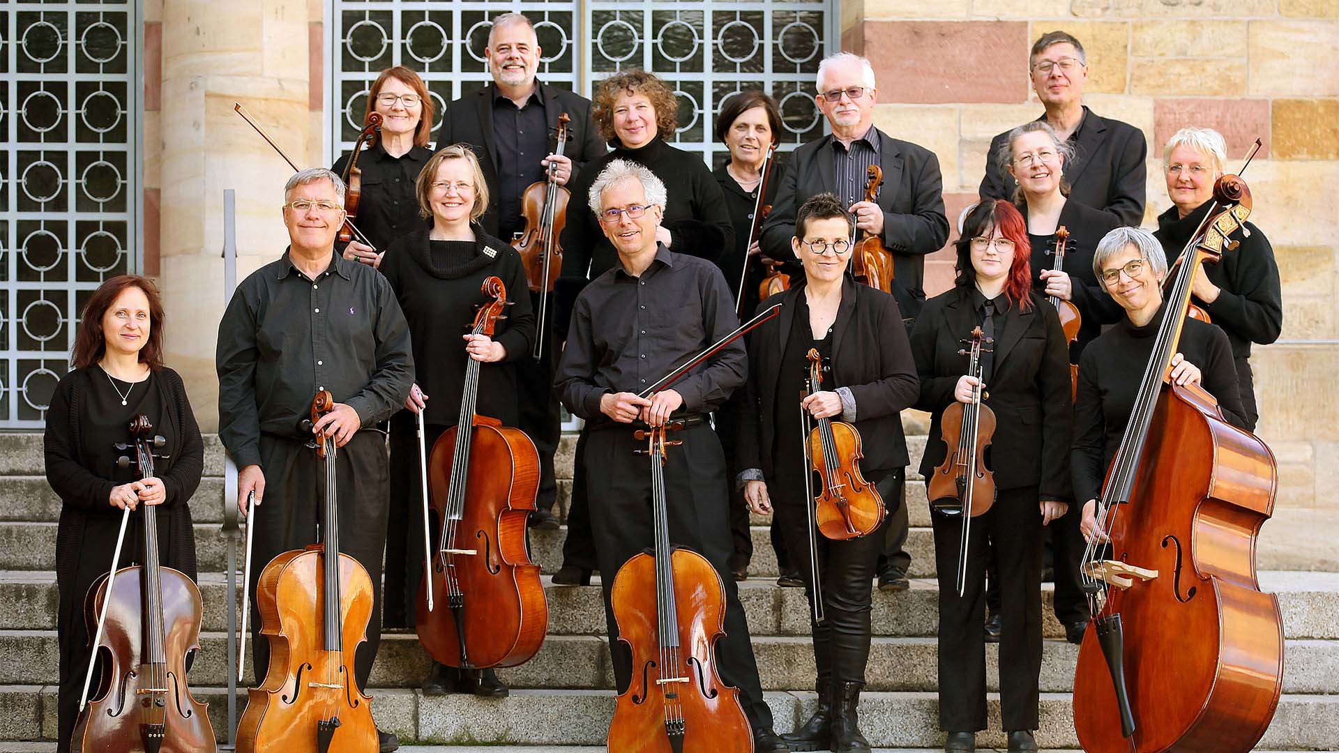 15 Musikerinnen und Musiker eines Streichorchesters stehen mit Violinen, Bratschen, Celli und Kontrabass auf einer Treppe vor einem Gebäude.