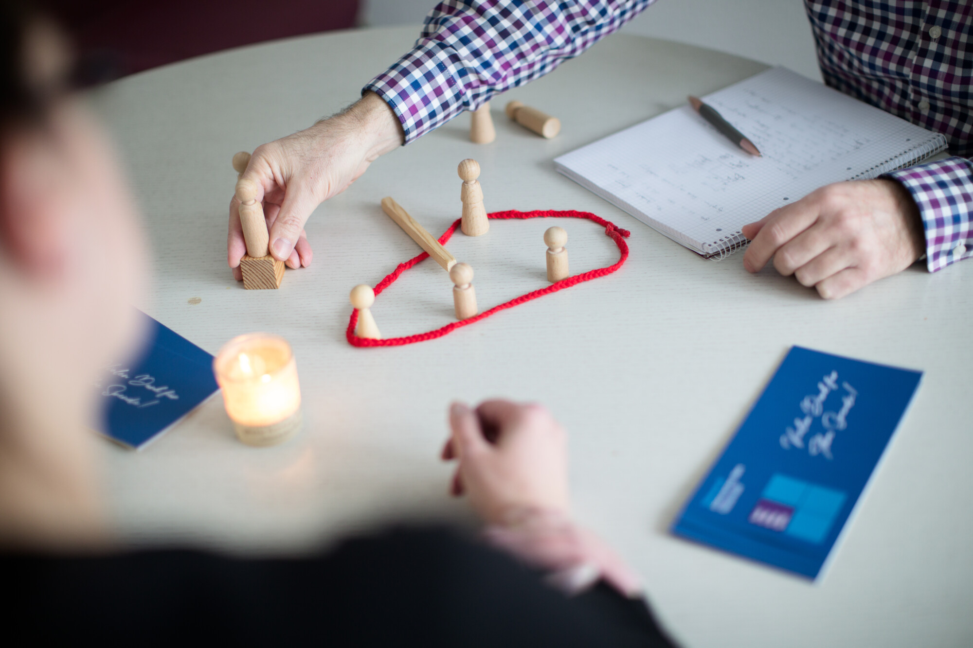 Blick auf einen Tisch, man sieht Hände von zwei Personen, eine Kerze, Flyer, einen Schreibblock mit Stift und Holzkegel, die innerhalb einer zum Kreis geschlossenen roten Schnur platziert werden.