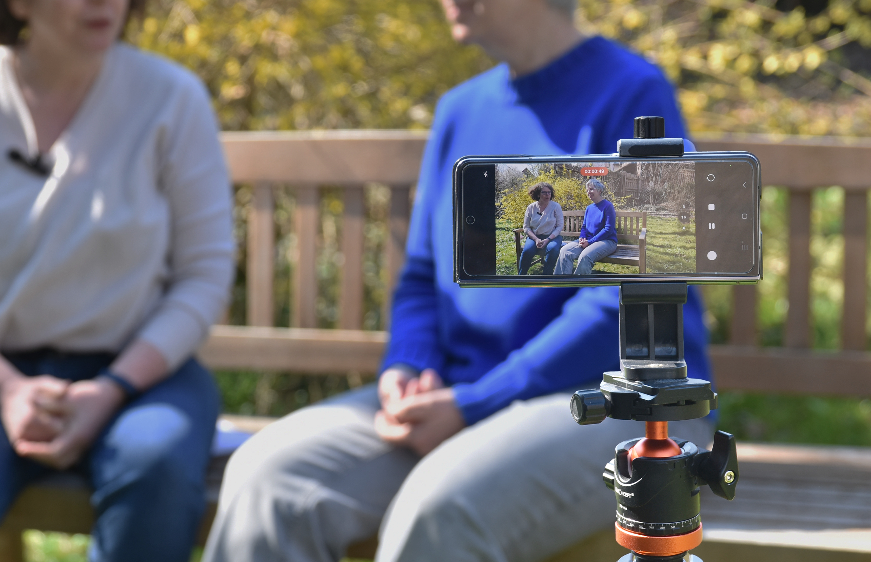 Kerstin Hillgärtner und Regine Jünger sitzen auf einer Bank im Garten, ihre Köpfe sieht man nicht. Dafür ein Stativ mit einem Smartphone. Im Display sieht man die Gesichter der beiden Pfarrerinnen.