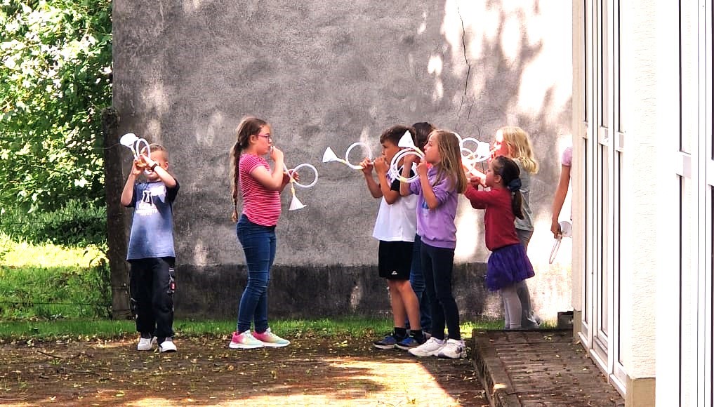 Kinderkirchenzeit Kinder blasen auf selbstgebastelten Trompeten an einer Mauer