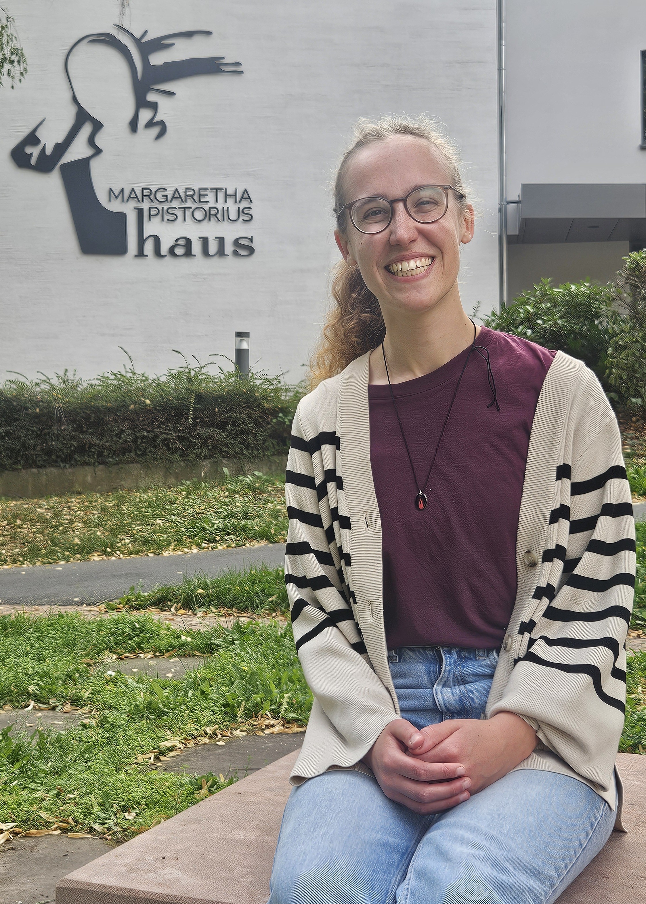 Lara Weller sitzt auf einem Sandstein-Quader im Johanniter-Park, im Hintergrund sieht man das Dekanatsgebäude und erkennt den Schriftzug Margaretha-Pistorius-Haus mit der abstrakten Darstellung der Margaretha-Pistorius. Lara Weller trägt Jeans, T-Shirt und Cardigan. Das lockige blonde Haar hat sie zum Zopf gebunden. Sie trägt eine Brille und lacht offen und fröhlich in die Kamera.