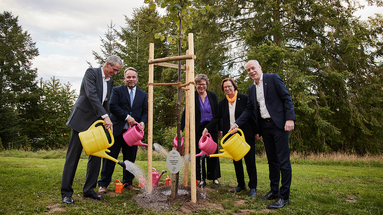 Menschen gießen mit bunten Gießkannen einen jungen Baum