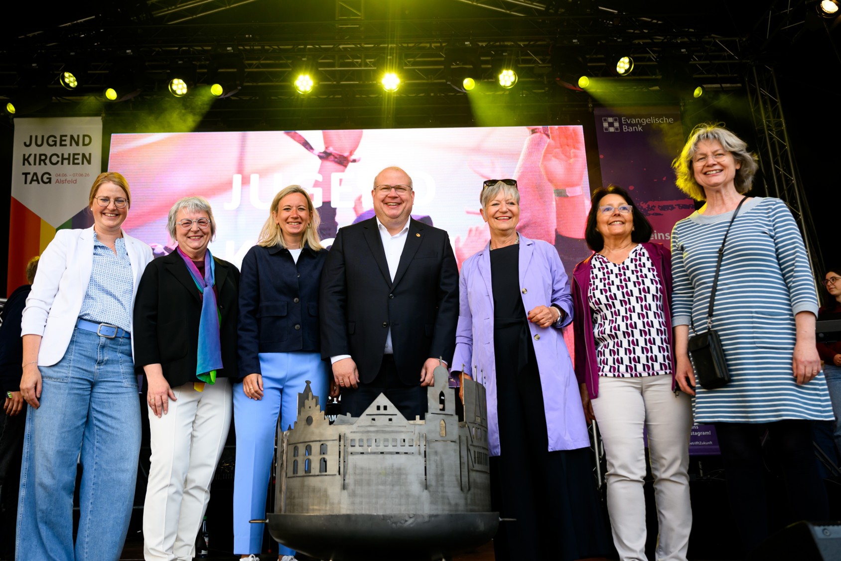 Sieben Erwachsene stehen nebeneinander auf einer Veranstaltungsbühne. Hinter ihnen sind eine große LED-Leinwand sowie Banner mit der Aufschrift „Jugendkirchentag“ und „Evangelische Bank“ zu sehen. Im Vordergrund befindet sich ein metallisches Modell eines Gebäudekomplexes. Bühnenbeleuchtung in Gelb und Grün beleuchtet die Szene.