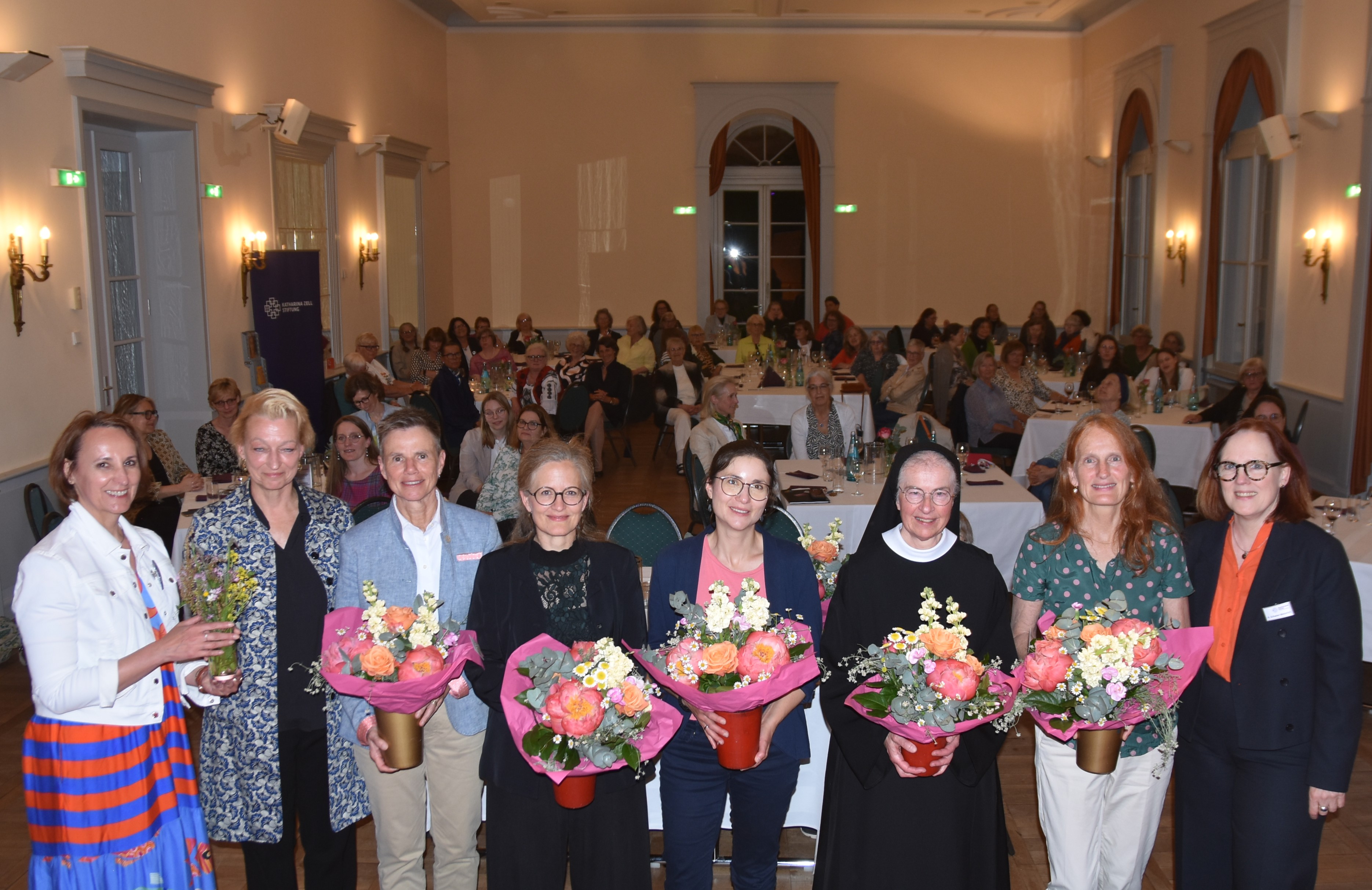 Acht Frauen stehen in einer Reihe nebeneinander. Sie sind - bis auf eine Nonne, bunt gekleidet, fünf Frauen halten einen großen Blumenstrauß in der Hand. Sie posieren für ein Gruppenfoto. Im Hintergrund sieht man, an Tischen sitzend, die weiblichen Gäste der Veranstaltung.