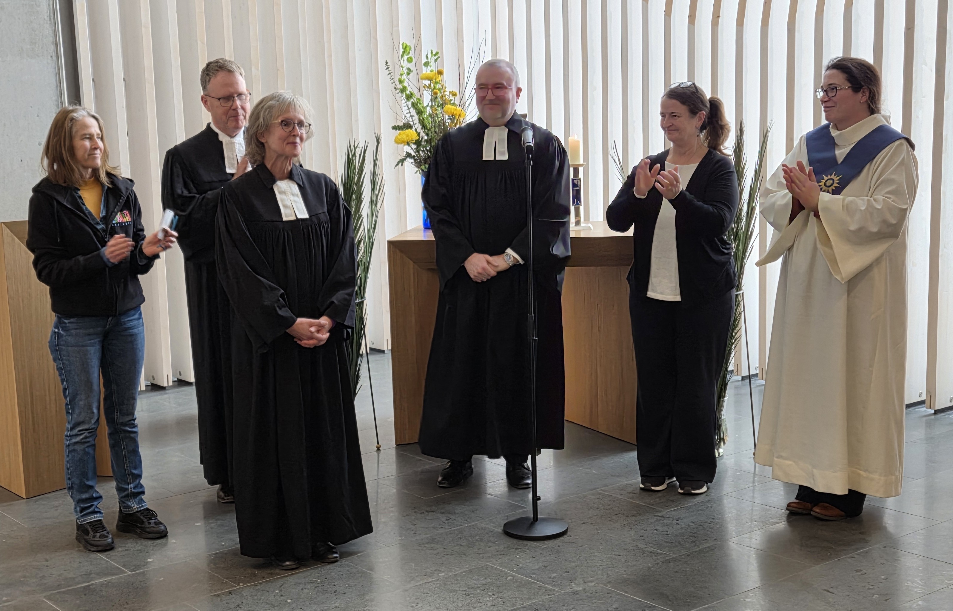Fünf Menschen stehen im Halbkreis hinter der Pfarrerin und klatschen Beifall