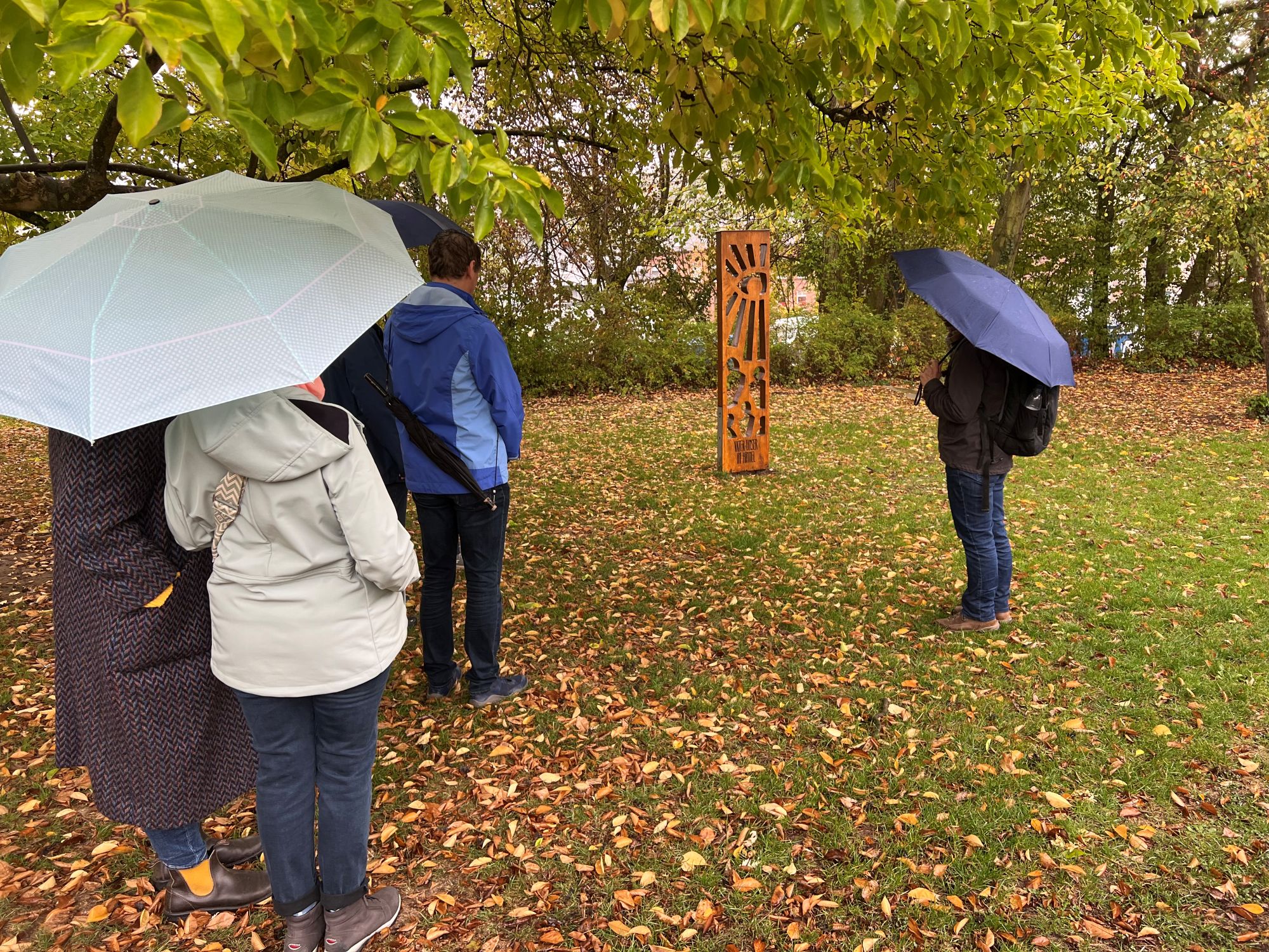 Menschen mit Regenschirmen stehen in einem parkähnlichen Bereich und schauen auf eine Holzskulptur