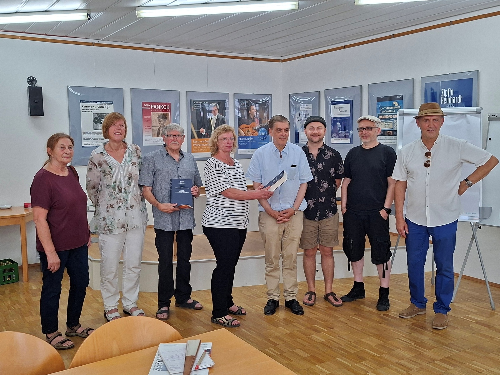 Neun Personen stehen in einer Reihe in einem hellen Raum mit Postern an der Wand und halten Bücher in den Händen. Langbeschreibung: Die Gruppe besteht aus sieben Männern und zwei Frauen, die in einem Raum mit Holzfußboden und Postern an der Wand stehen. Einige tragen lässige Kleidung wie T-Shirts, Hemden und Hosen, andere haben Accessoires wie Hüte oder Brillen. Zwei Personen in der Mitte halten Bücher, die sie präsentieren. Im Hintergrund sind Stühle und ein Tisch mit Materialien zu sehen.