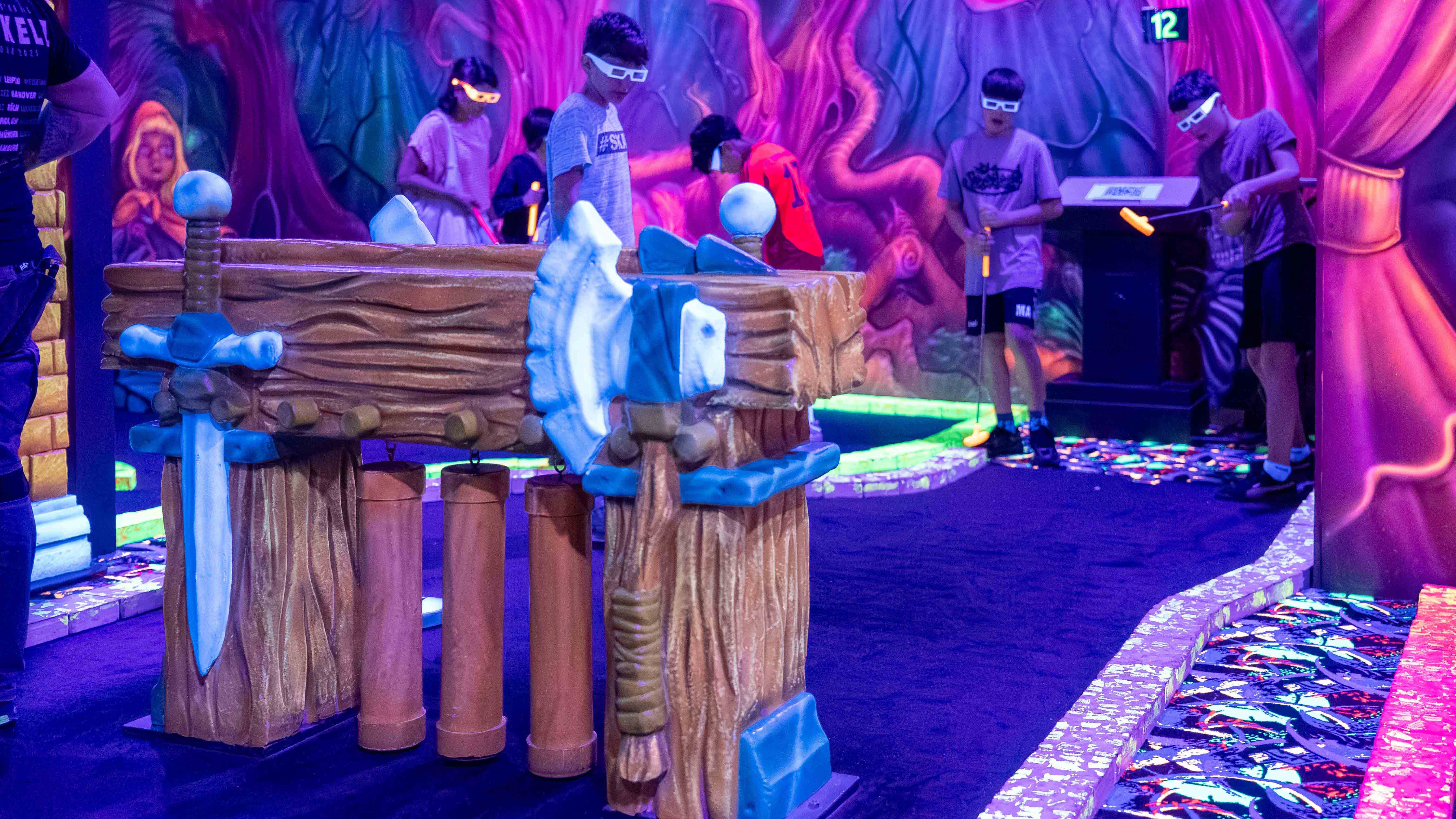 Kinder spielen Minigolf in einer Indoor-Anlage mit Schwarzlicht, bunten Wandmotiven und leuchtenden Schlägern. 