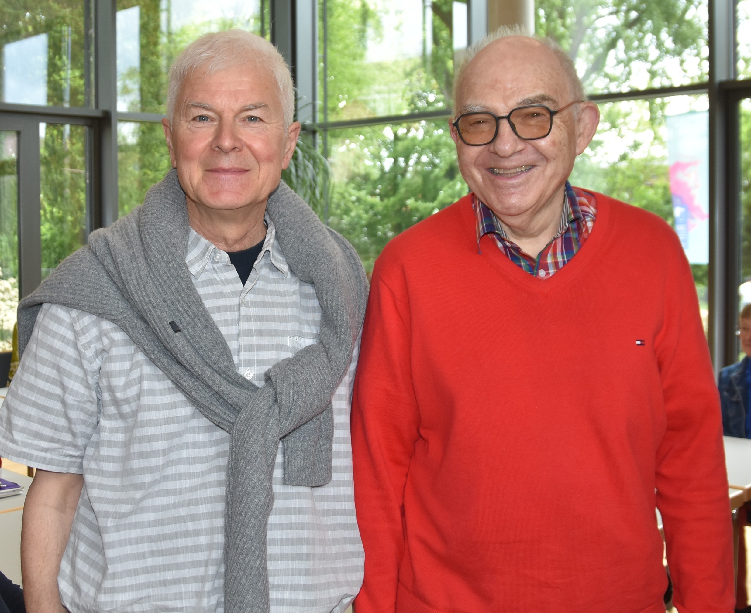 Gerhard Griestock, kurzärmliges Hemd und über die Schultern gelegter Pullover, und Rolf Hartmann, roter Pullover, blaues Poloshirt, stehen nebeneinander im Johannitersaal des Dekanats und lächeln in die Kamera