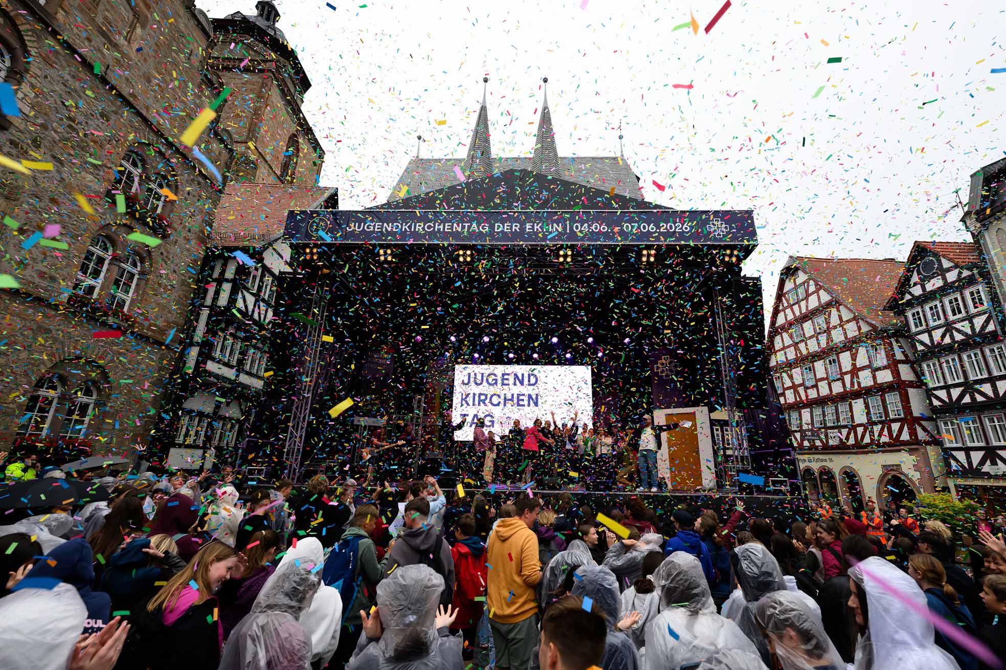 Auf einem Marktplatz mit Fachwerkhäusern und einer Kirche im Hintergrund steht eine große Bühne. Auf Bildschirmen und Bannern ist „Jugendkirchentag“ zu lesen. Viele Menschen versammeln sich vor der Bühne, einige tragen Regenponchos. Während Personen auf der Bühne stehen, regnet buntes Konfetti über die gesamte Szene.
