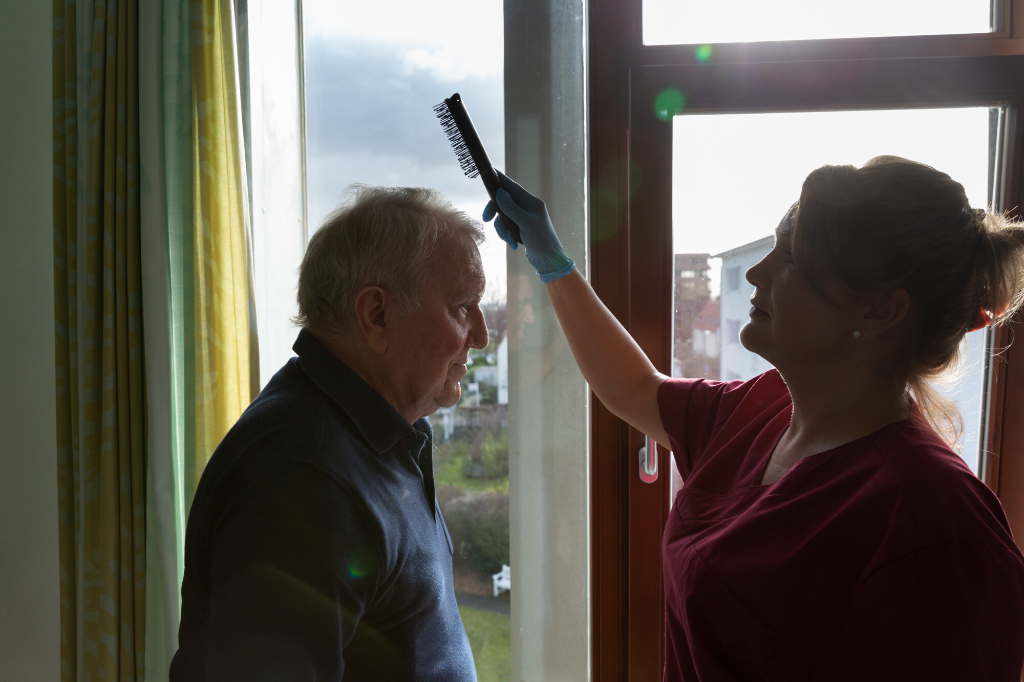 Aufnahme im Gegenlicht, Personen hauptsächlich als Konturen zu erkennen. Links steht ein alter Mann, ihm gegenüber eine jüngere Frau, die einen Kamm in der rechten Hand hält und ansetzt, um den Mann zu kämmen.