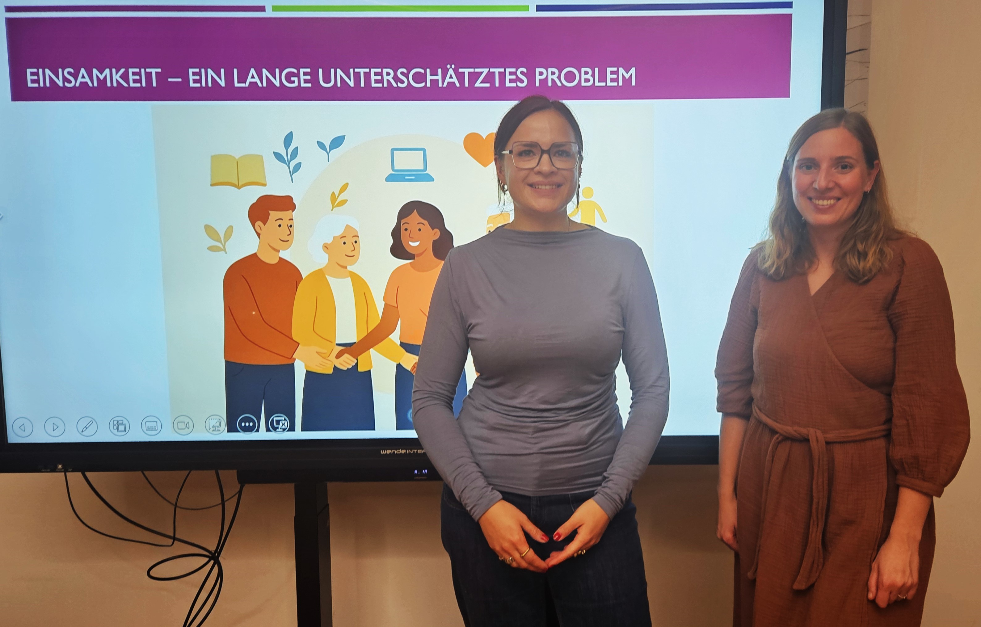 Maria-Louise Seipel (links) und Annelie Meis stehen vor einer Videowand, auf der eine PP-Folie zum Thema Einsamkeit zu erkennen ist, und lächeln in die Kamera.