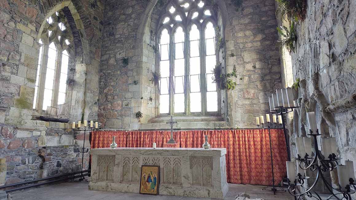 Der Altarraum einer alten Steinkirche oder Klosterruine. Hinter dem steinernen Altar befinden sich drei große, hohe Fenster mit gotischen Bögen. Mehrere Kerzenständer stehen rechts und links des Altars. Vor dem Altar lehnt ein kleines religiöses Gemälde. Die Steinwände zeigen Spuren von Alterung und vereinzelten Pflanzenbewuchs.