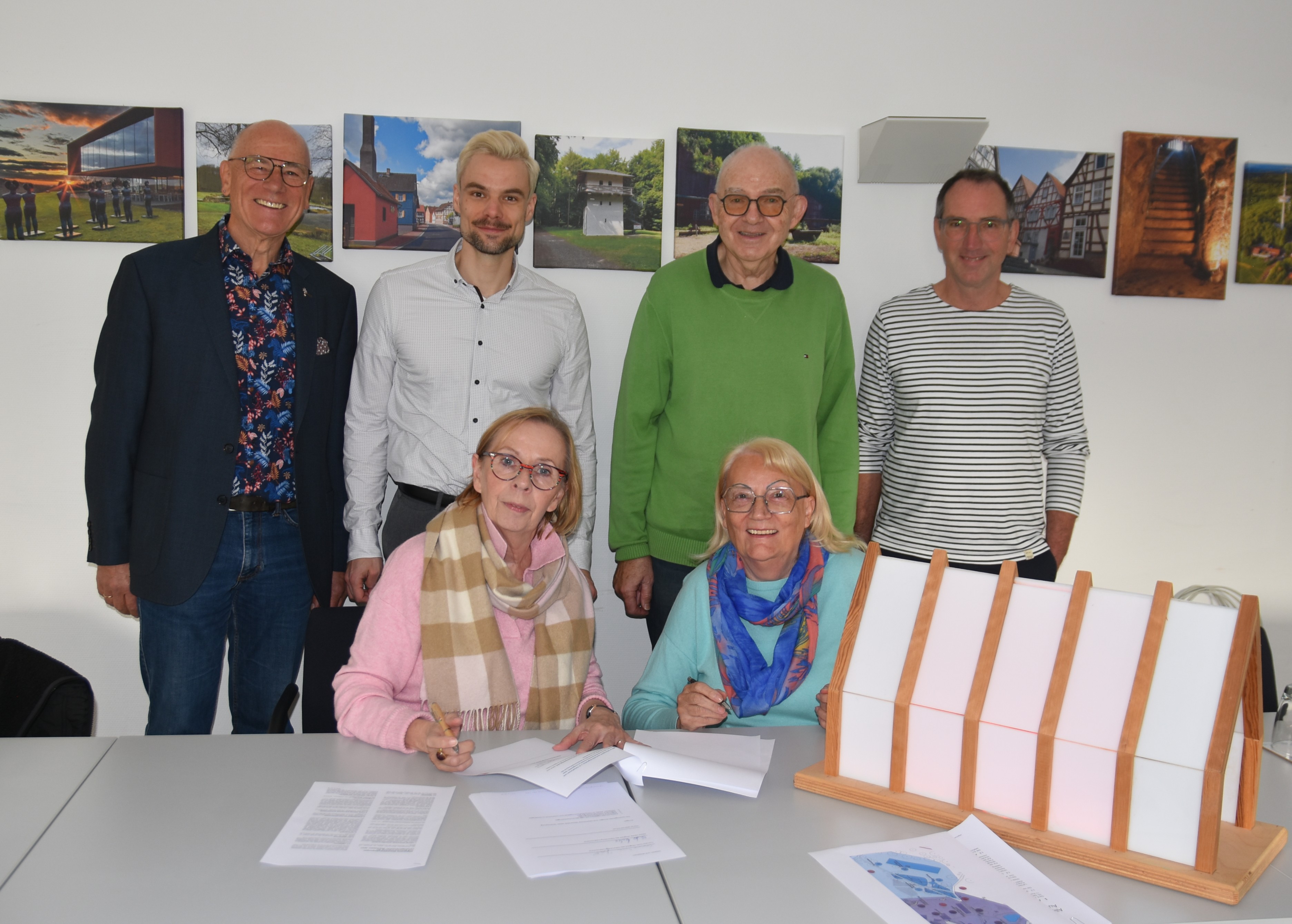 Sechs Personen, zwei Frauen sitzend an einem Tisch, vor sich Papiere, die sie unterschreiben, dahinter stehen sechs Männer. Auf dem Tisch steht ein Modell der Lichtkirche. An der Wand hinter den Männern hängen Fotografien.