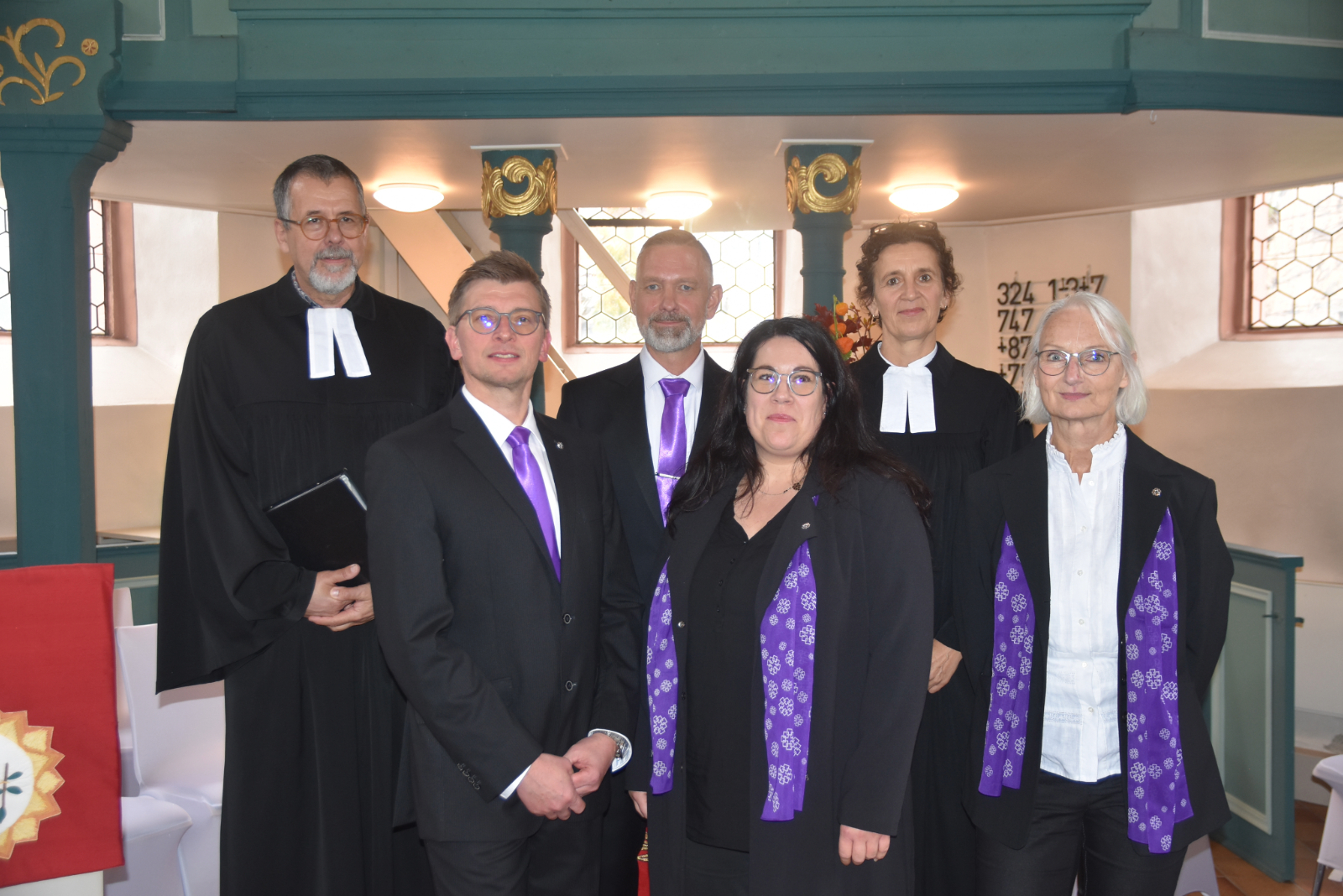 Pröpstin Anke Spory und Pfarrer Jürgen Füg, beide im Talar, mit dem vierköpfigen Bergheimer Kirchenvorstand, der schwarz und lila gekleidet ist.