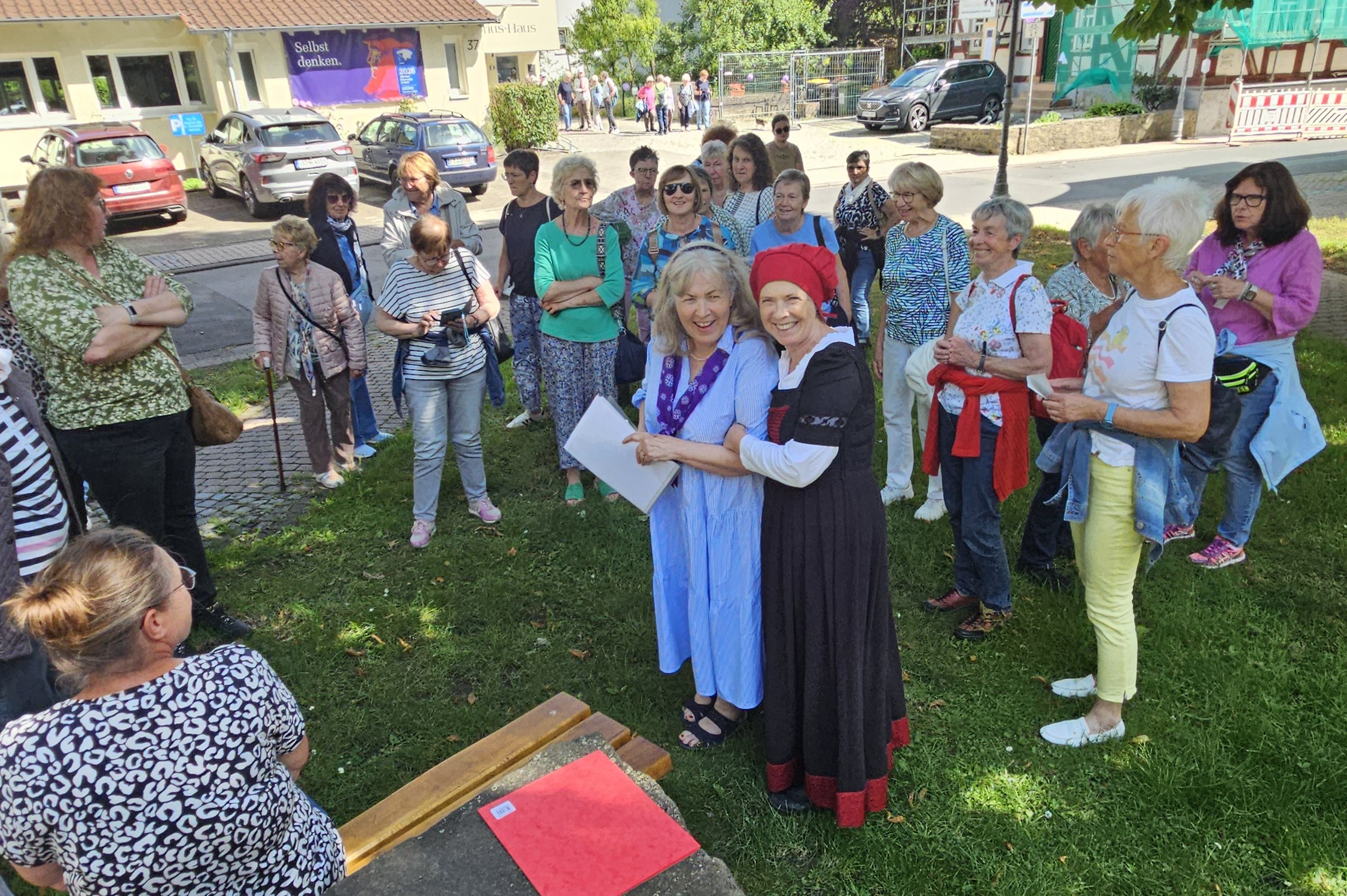 Eine große Gruppe von Frauen auf einer Wiese steht um Beate Harbich-Schönert und Andrea Jungnick-Hoeltgen, die sich umschlungen halten. Beate Harbich-Schönert trägt ein hellblaues Sommerkleid, Andrea Jungnick-Hoeltgen ist in der Tracht einer Pfarrfrau im 16. Jahrhundert.