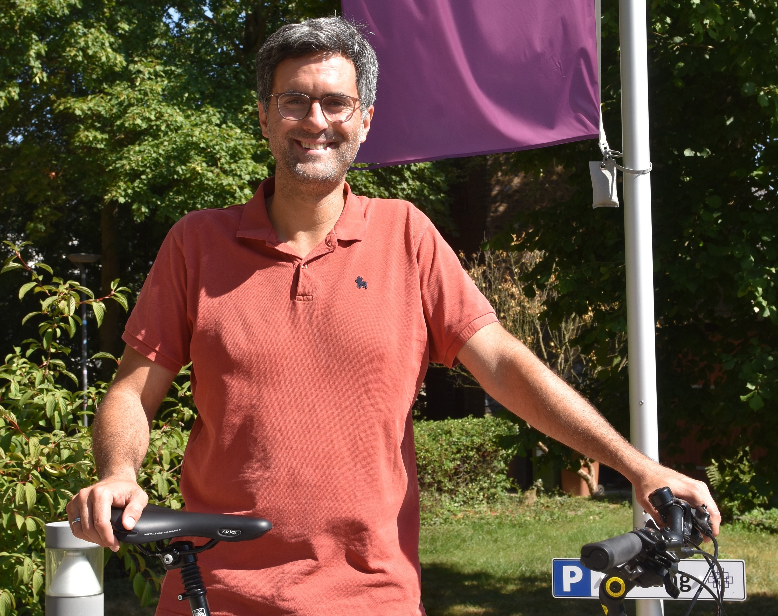 Andreas Hlasseck, kurze dunkle Haare, Brille, rotes Poloshirt, steht vor einer lilafarbenen EKHN-Fahne, von der man nur einen Teil sieht, vor ihm sein Fahrrad, das er an Sattel und Lenker festhält. Im Hintergrund grüne Hecken und Bäume.
