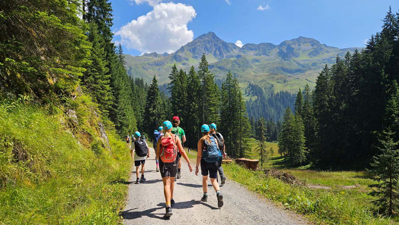 Gruppe von Kindern und Jugendlichen mit Rucksäcken wandert auf einem Bergweg durch einen Nadelwald, im Hintergrund hohe Berge und blauer Himmel.