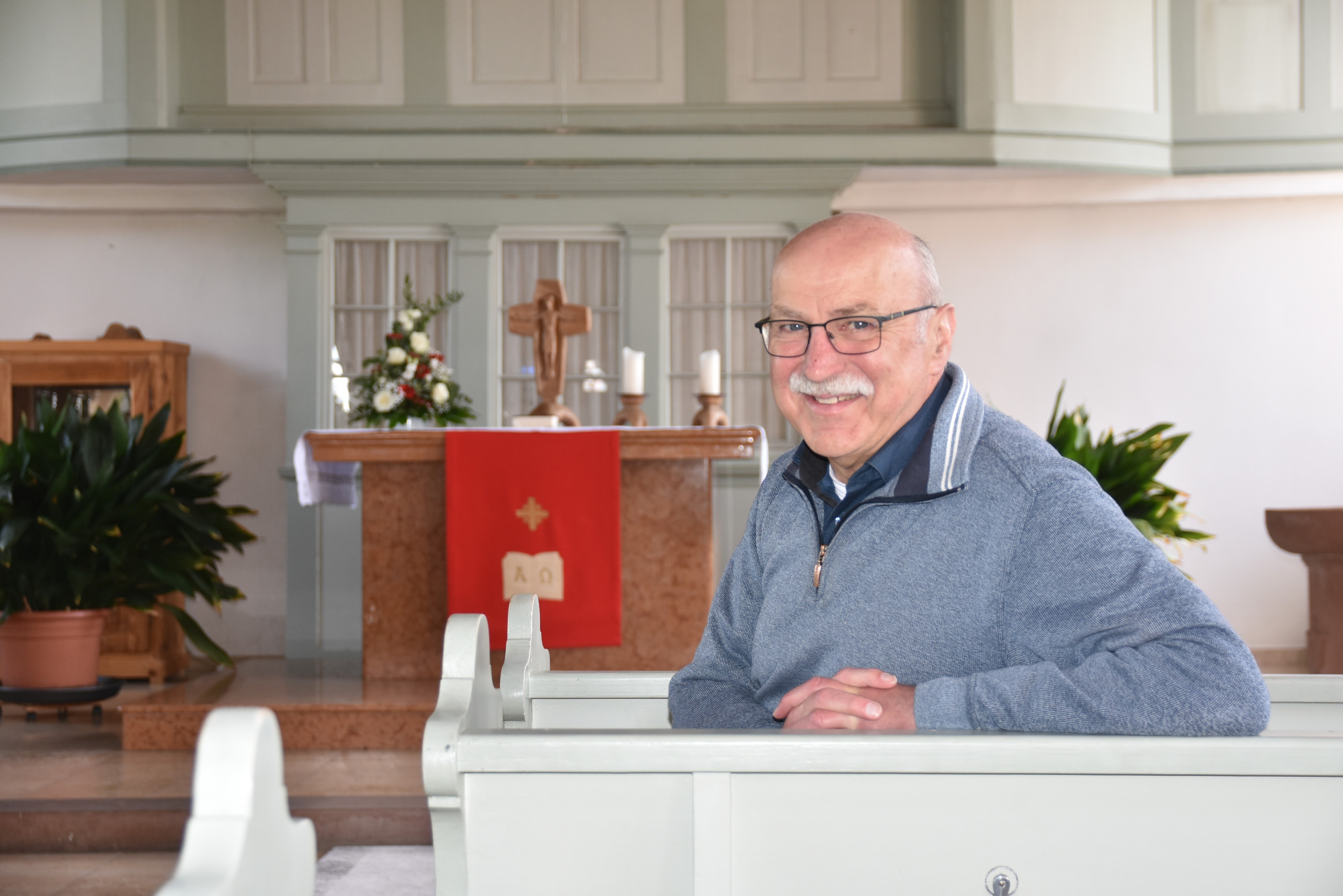 Pfarrer Dieter Wichihowski sitzt in der Oberauer Kirche auf einer Bank mit dem Rücken zum Alter und hat die Arme auf der Rückenlehne abgelegt. Er lächelt in die Kamera. Im Hintergrund sieht man den Altar mit Kerzen, Blumenschmuck und einer roten Altardecke und lächelt in die Kamera. Er trägt Hemd und Pullover. Im Hintergrund sieht man den 