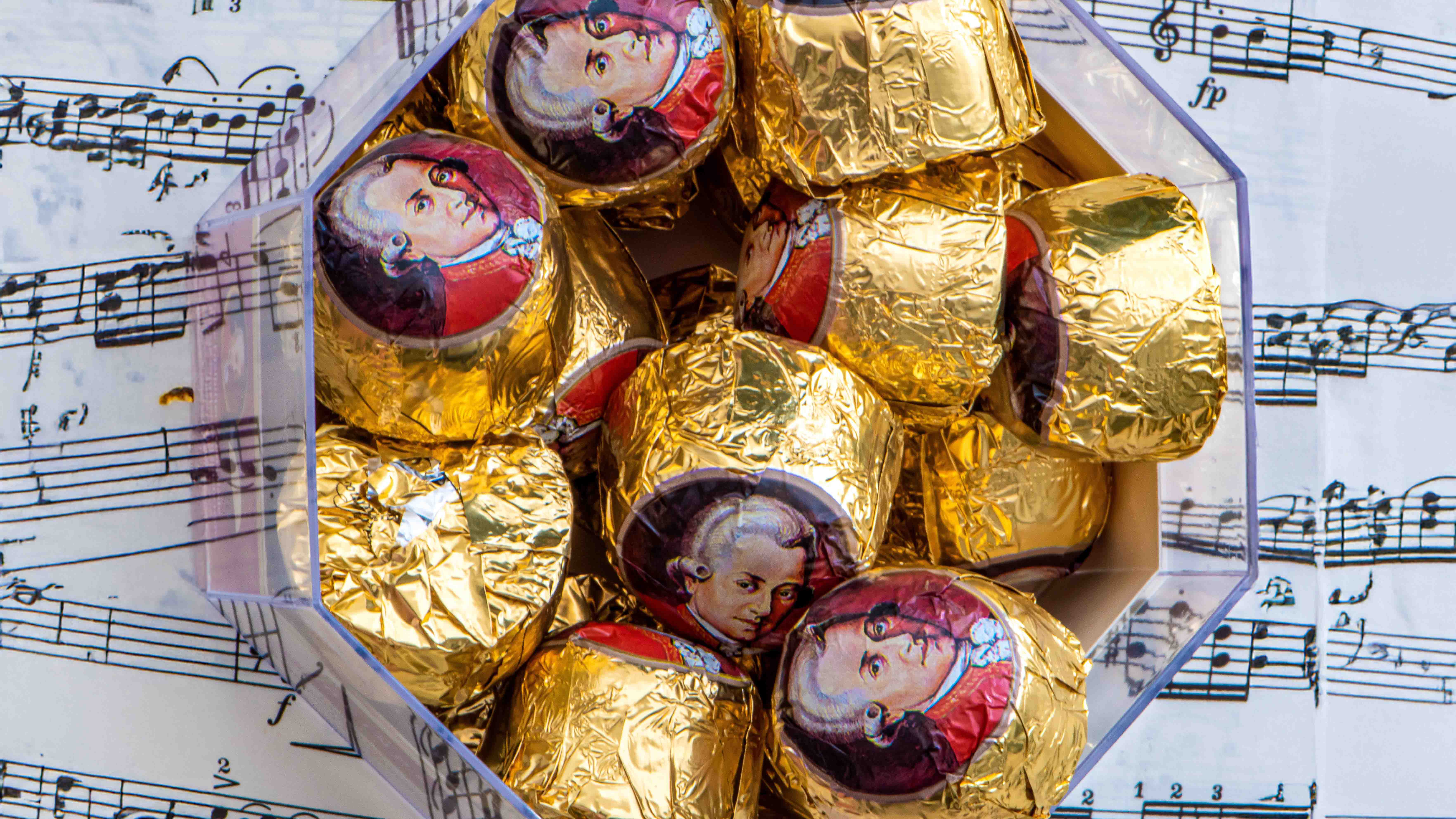 Goldverpackte Mozartkugeln mit Mozart-Porträts liegen in einer Schachtel auf ausgebreiteten Notenblättern.