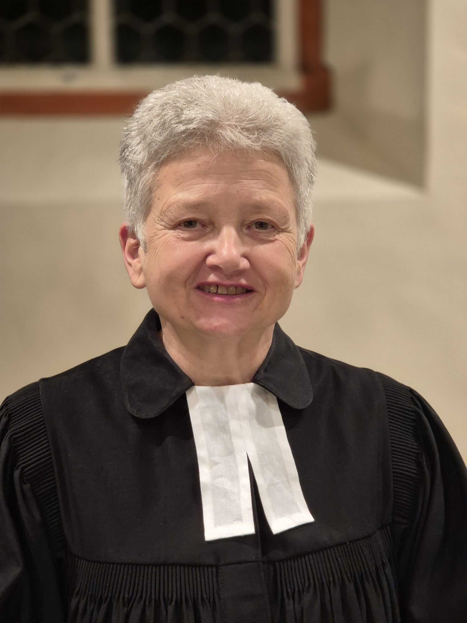 Porträt Andrea Klimm-Haag im Talar in einem Kirchenraum. Sie hat sehr kurze graue Haare und blickt freundlich in die Kamera.