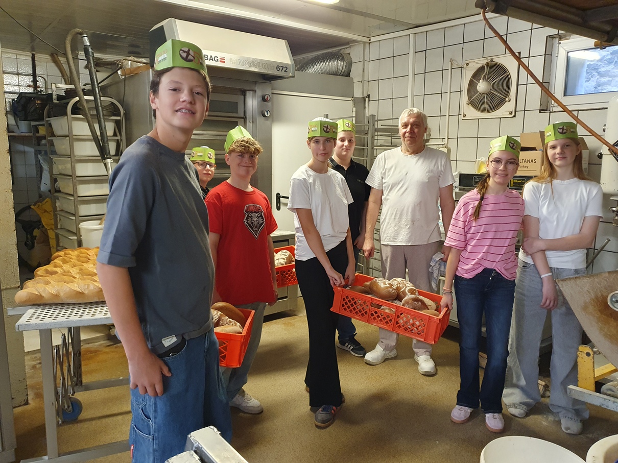 Sieben Personen in einer Bäckerei, sechs mit grünen Brotzeit-Hüten, vor Backöfen und einem Tisch mit langen Brotlaiben; eine Person hält einen roten Korb mit Broten.