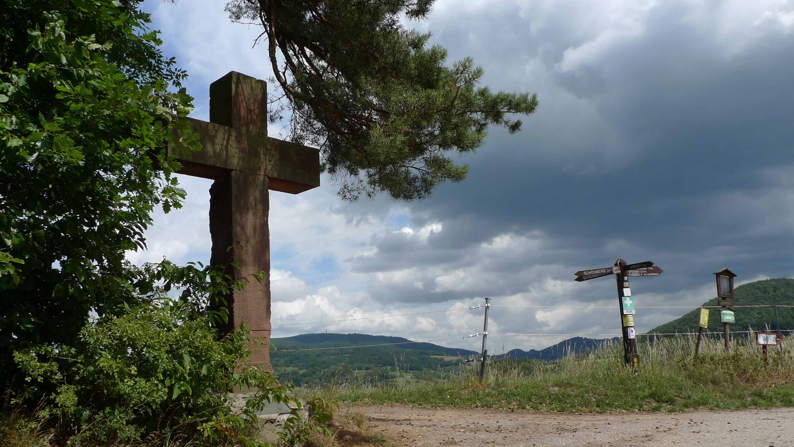 Großes steinernes Kreuz am Waldrand, daneben Wanderwegweiser vor einer hügeligen Landschaft unter dunklen Wolken.