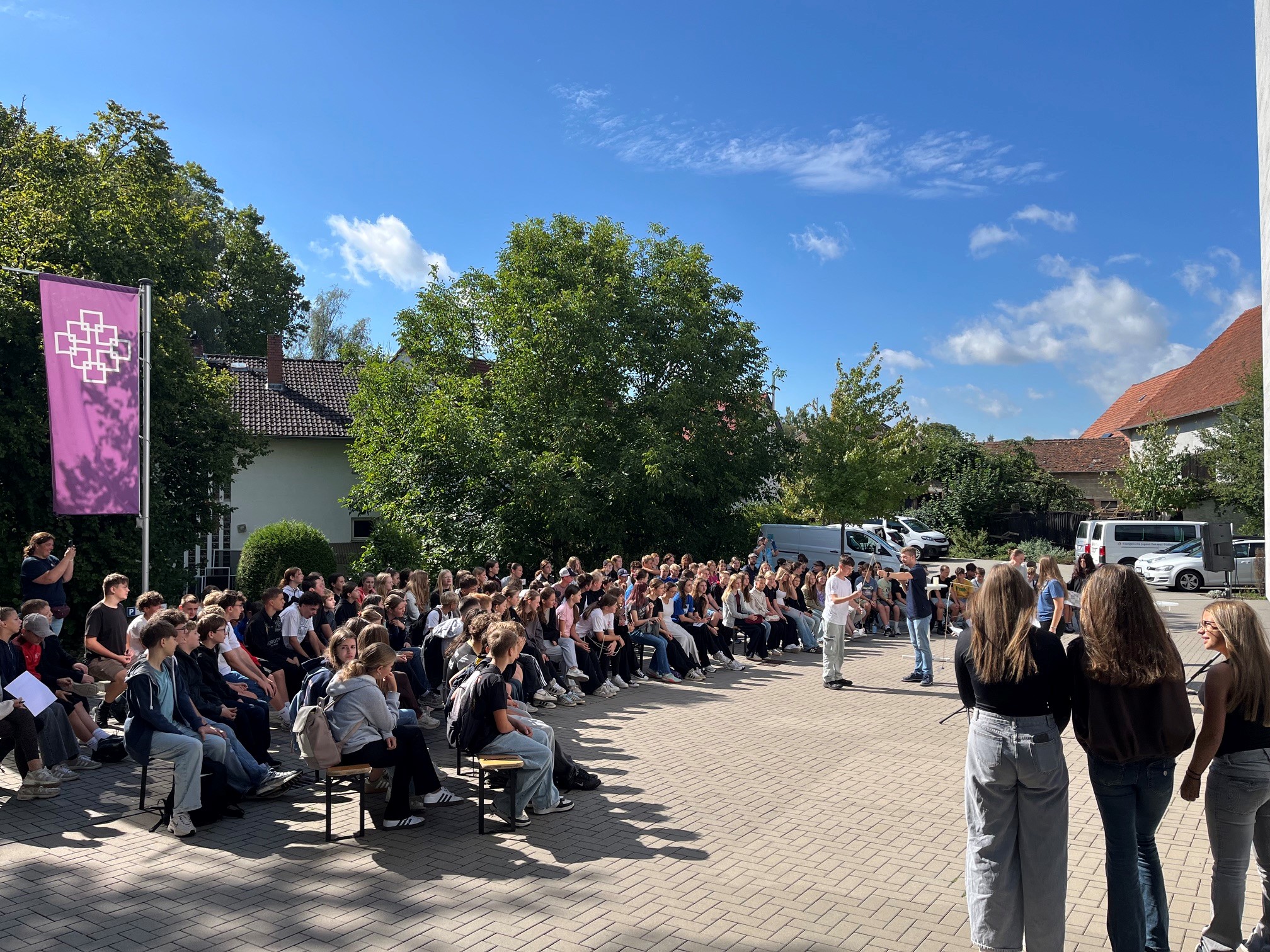 Blauer Himmel mit ein paar Wolken, Blick auf den Parkplatz des Margaretha-Pistorius-Hauses. Auf Bänken sitzen sehr viele Konfirmandinnen und Konfirmanden, im Vordergrund stehen drei Personen mit dem Rücken zum Betrachter. In der Mitte des Platzes steht jemand und spricht zu den Konfirmandinnen und Konfirmanden. Links im Bild weht eine lila EKHN-Fahne.